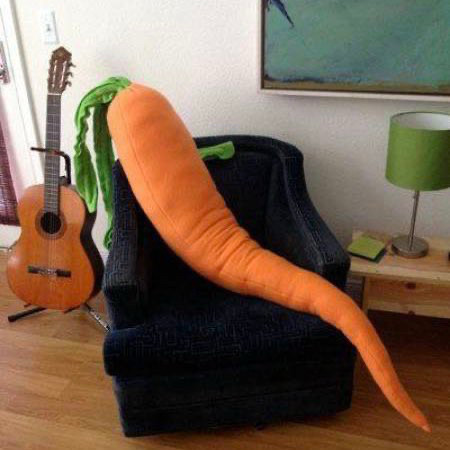 A giant stuffed carrot sits on a chair next to a side table with a lamp, and a guitar on a stand.