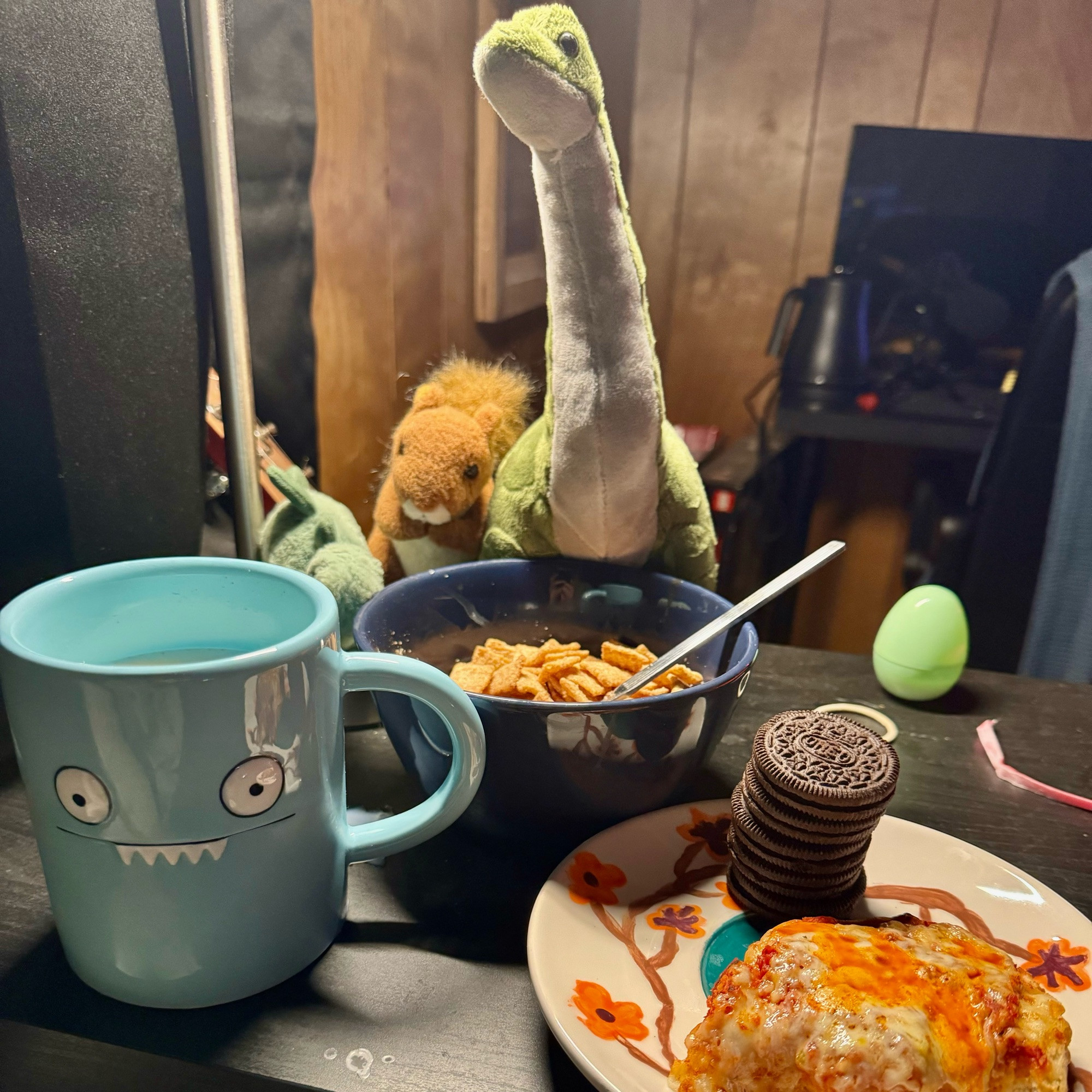 In order right to left, an “Icebat” mug fill with milk, a bowl of cinnamon toast crunch (kroger brand), and a plate containing a stack of oreos and a half pizza bagel