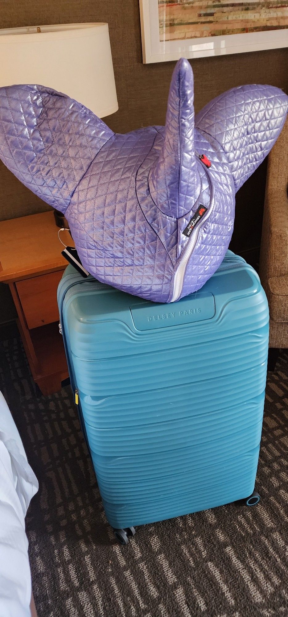 A purple suspiciously unicorn shaped fursuit head bag sits on top of a teal hardside suitcase. The con is over.