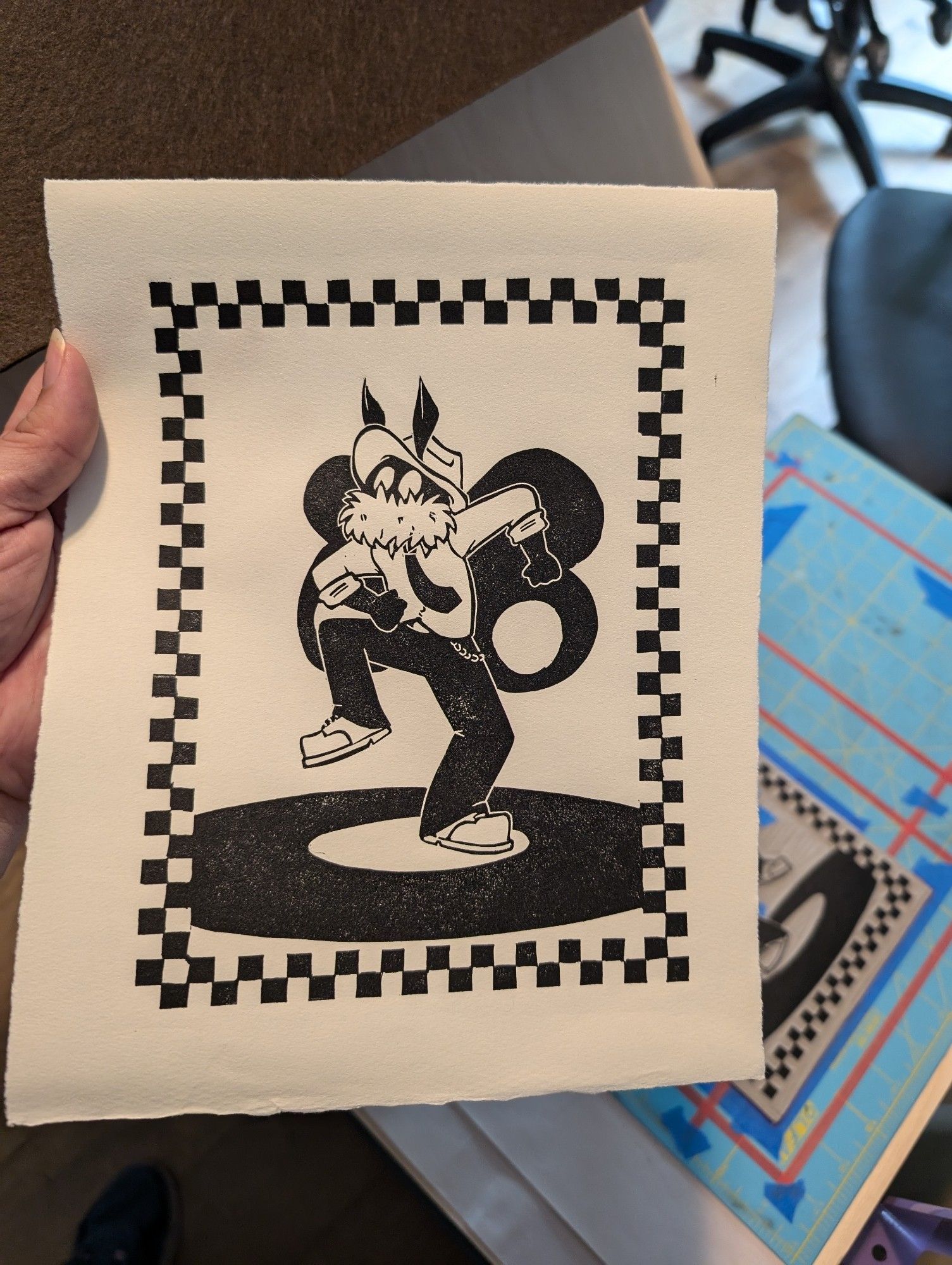 photo of a freshly printed block print. Mothman, dressed in black slacks, white shirt, black tie, fedora and boots, has his fists thrown to one side and one knew in the air while he skanks (it's a ska dance) on top of a record. the print is surrounded by a black and white checkered border. behind the print, the press, lino block, and registration mat can be seen.