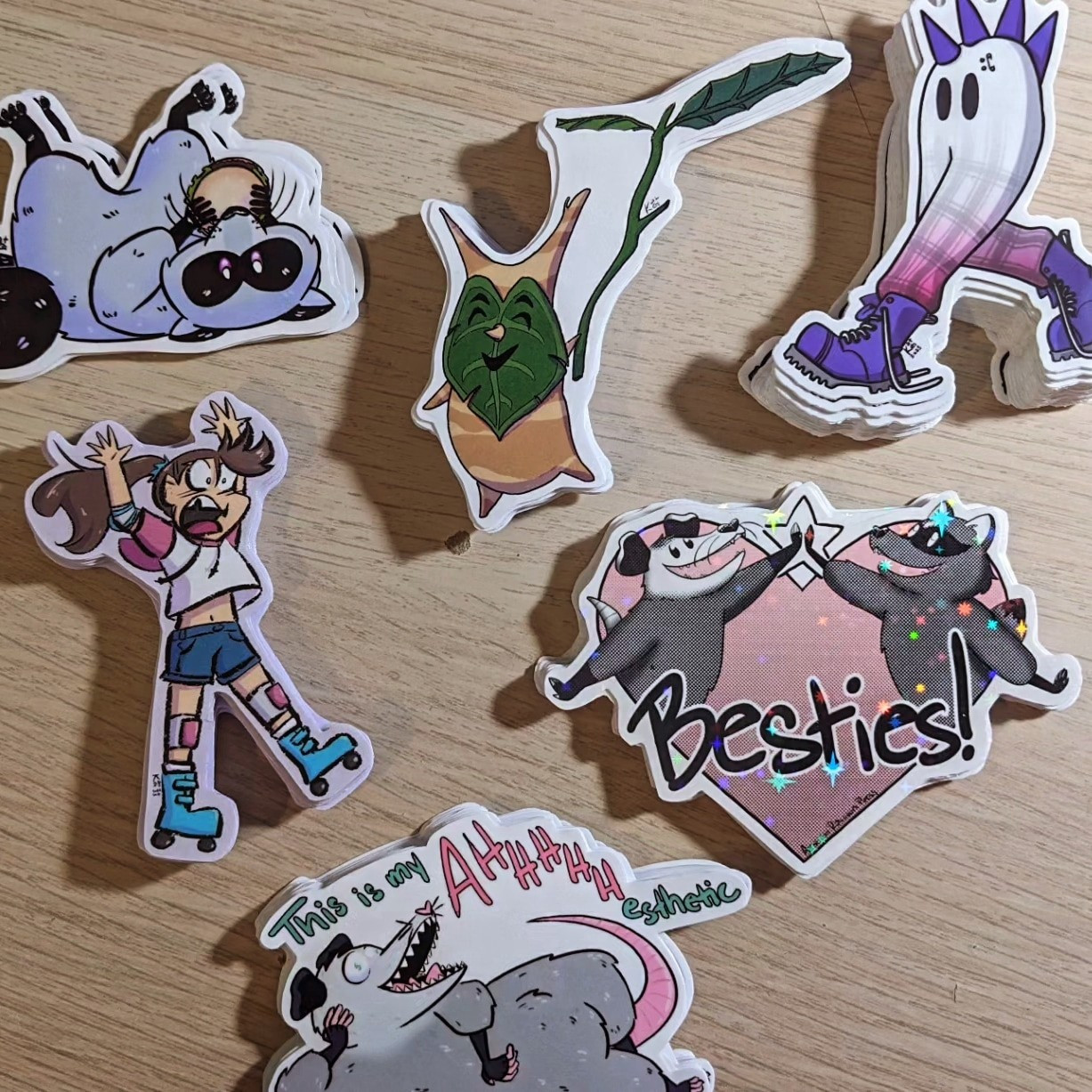 a photo of 6 piles of stickers. the stickers are of a raccoon lying on its back eating a hamburger, a korok, a person with a pony tail failing to roller skate, a punked out fresno nightcrawler, an oppossum and a raccoon high fiving in a heart with the word "besties!", and an oppossom lying like burt reynolds did on that bearskin rug and the words "this is my AHHHHHesthethic"