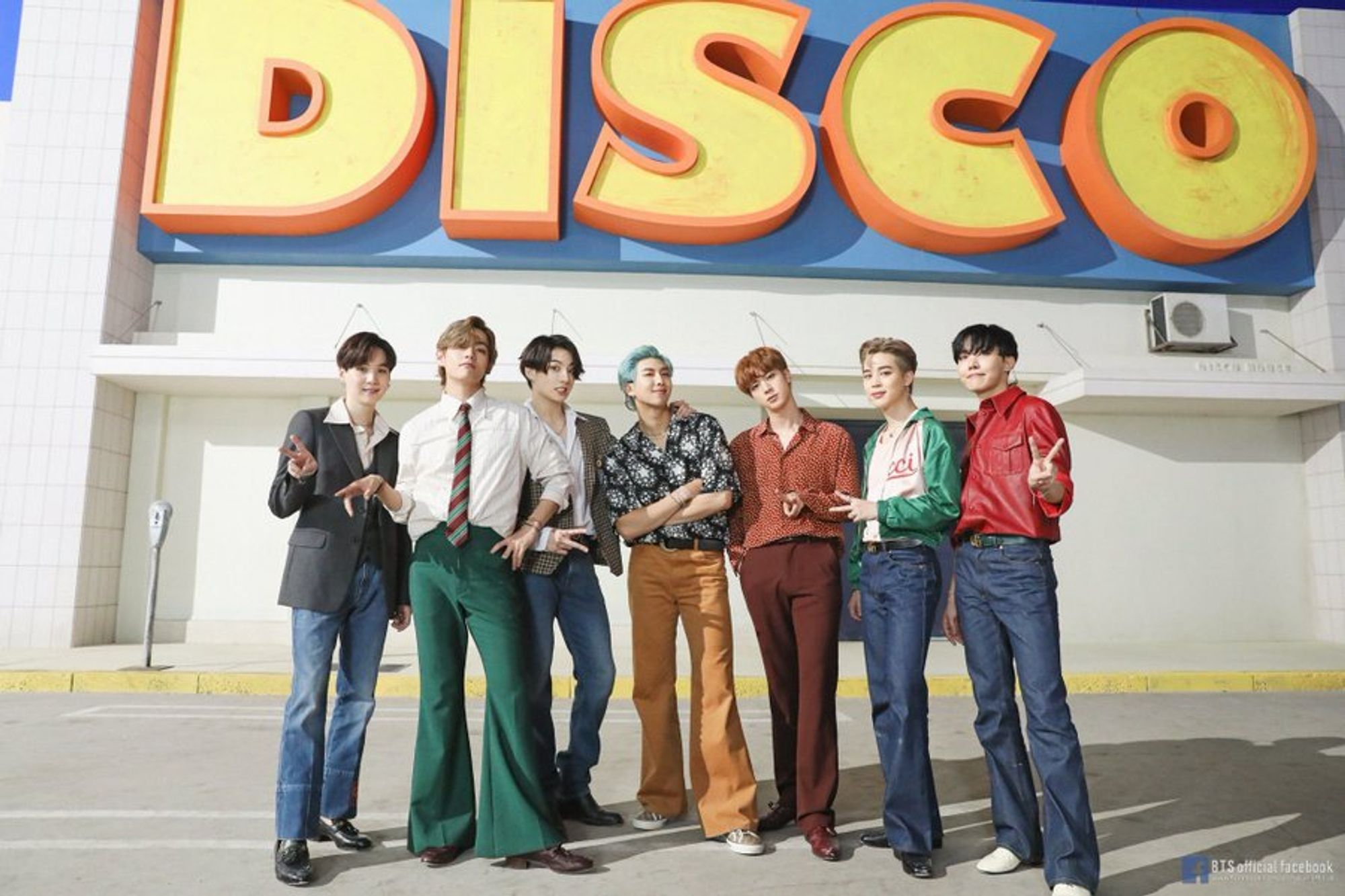 Picture of BTS on the set of the Dynamite music video, in front of a big sign that says DISCO, wearing 70s inspired clothing