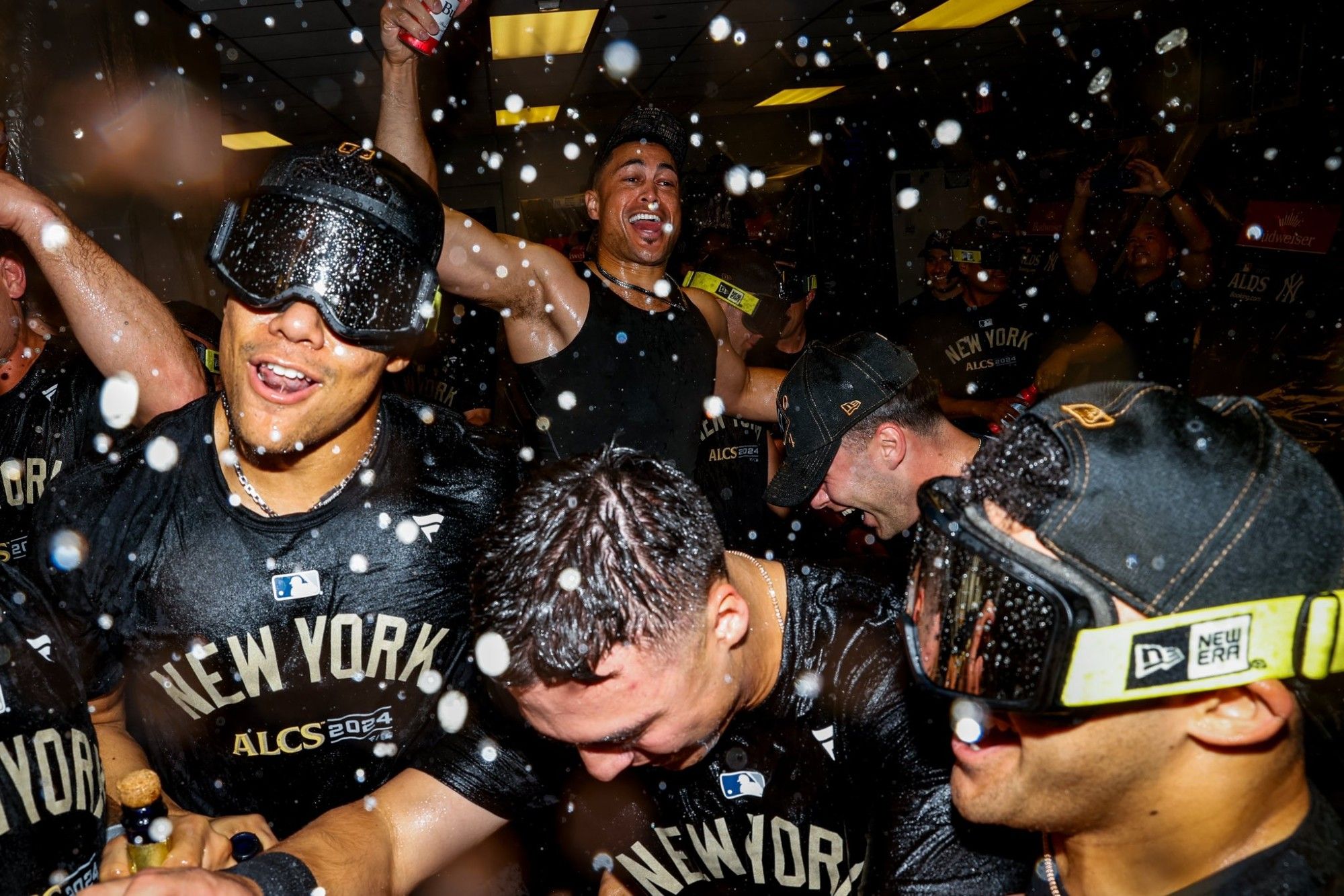 Giancarlo Stanton jumping around in the background while Juan Soto, Anthony Volpe, and Jasson Dominguez spray champagne closer to the camera.