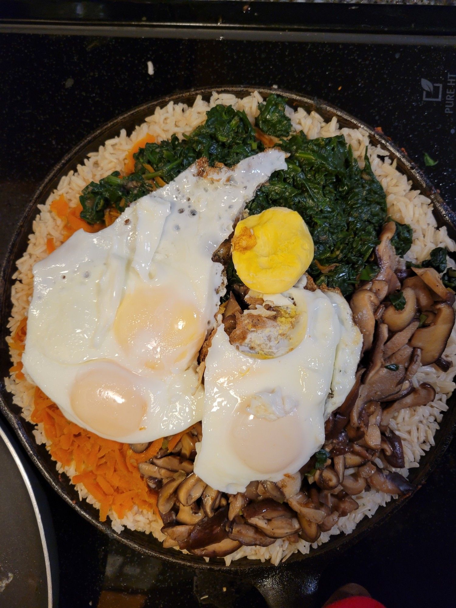 Bibimbap, a Korean rice bowl, on top of a stove. On top of rice are cooked mushrooms, carrots, spinach, and 4 cooked eggs.  One of the yolks has broken out of one of the eggs, and has fallen down the slope a bit.