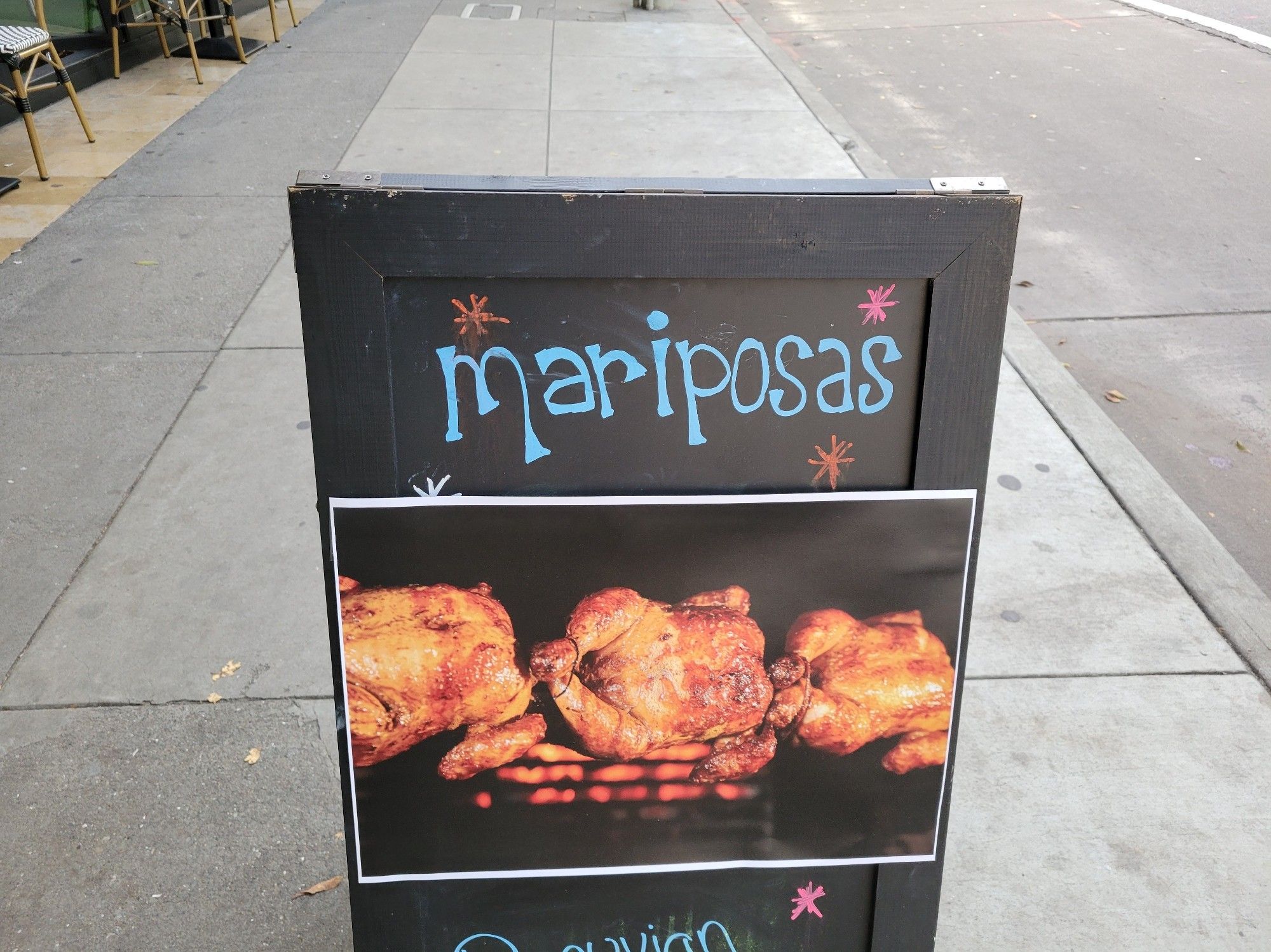 Sandwich board outside a restaurant with a photo of several chickens roasting on a spit, with the word "Mariposas" above them.