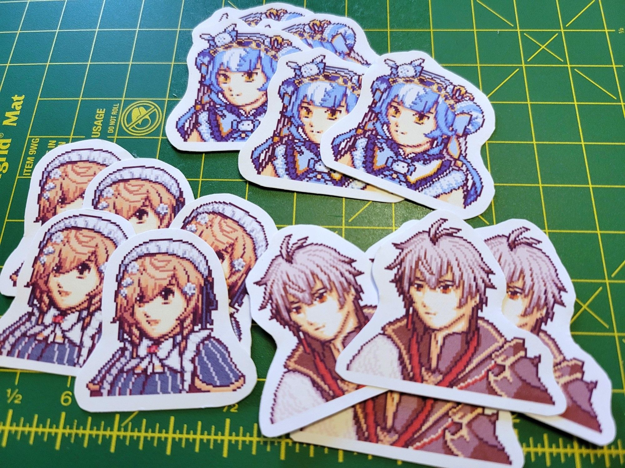 White bordered stickers of Nijisanji EN VTubers Twisty Amanozako, Klara Charmwood, and Ryoma Barrenwort on a green and yellow cutting mat. The characters are drawn in a pixelated style similar to the GBA Fire Emblem games