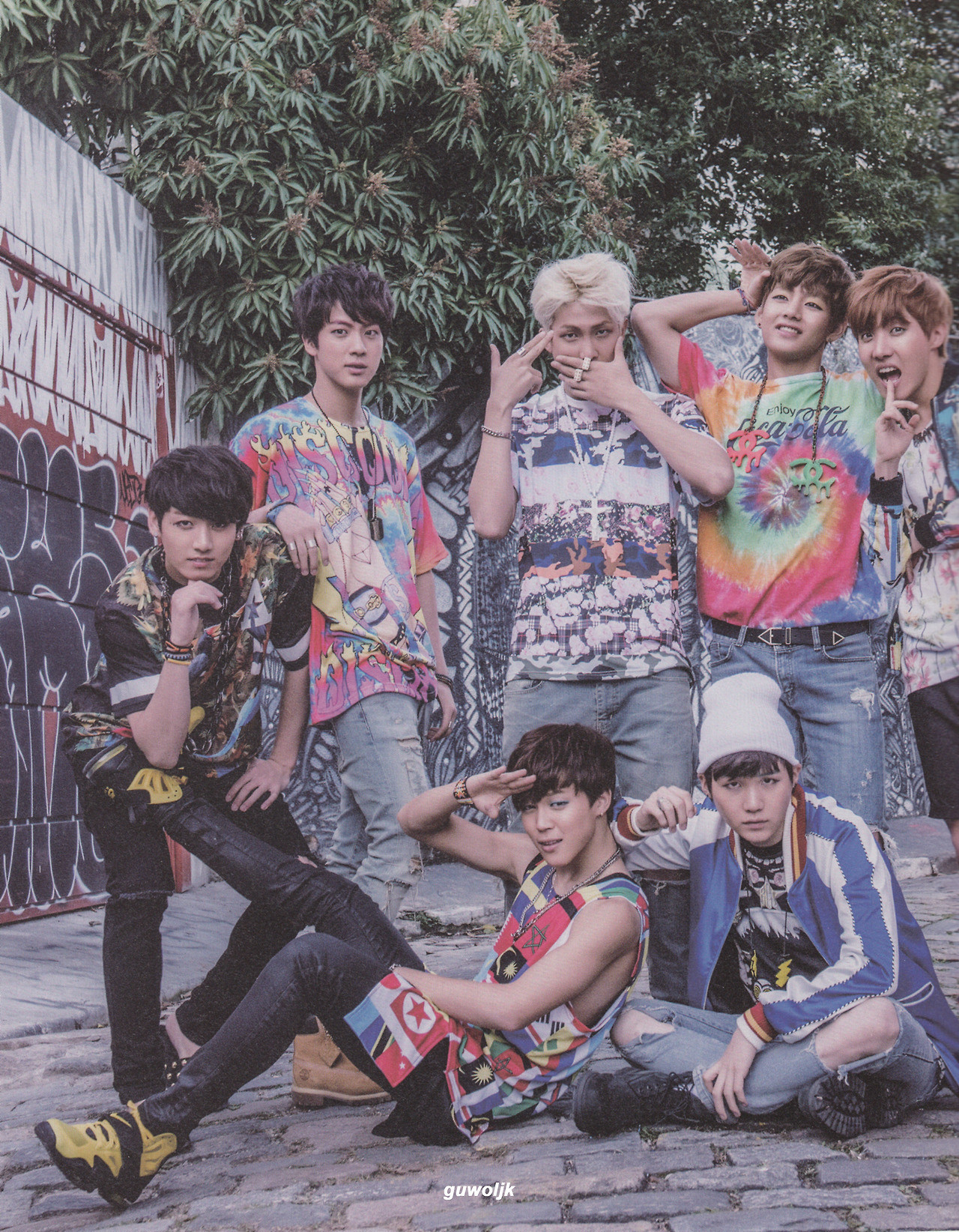 bts ot7 from their now2 photobook in 2015. they're dressed in very busy graphic or tie-dye shirts in rainbow colors and they're outside on a cobblestone street next to a wall covered in grafitti. they're all posing in crazy ways. hobi's finger is almost in his mouth, taehyung has one hand pressed to his head like a swooning victorian maiden, namjoon is covering his own mouth and pointing a finger gun at his own head. jungkook is leaned over with one elbow braced on his knee and his chin on his hand. jimin, who is sitting on the ground, has one leg raised so his foot is being held between jungkook's knee and elbow. he's also saluting? yoongi is next to jimin staring intensely into the camera. jin is standing there like some super handsome guy who just wandered into the shoot and is a little scared. they're all wearing lots of eyeliner, and some of them are also wearing pastel eyeshadow or face gems. inspired.