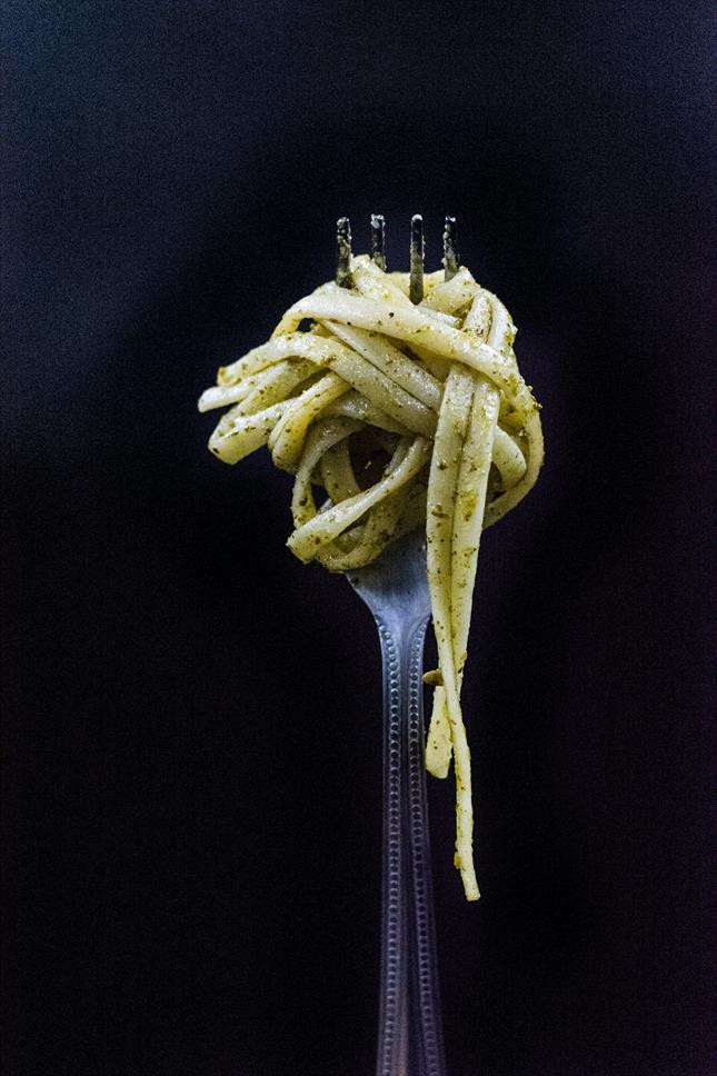 Color photo: a knot of pasta (pesto linguine?) spun around a silver fork viewed in extreme close up in front of a black-draped background.