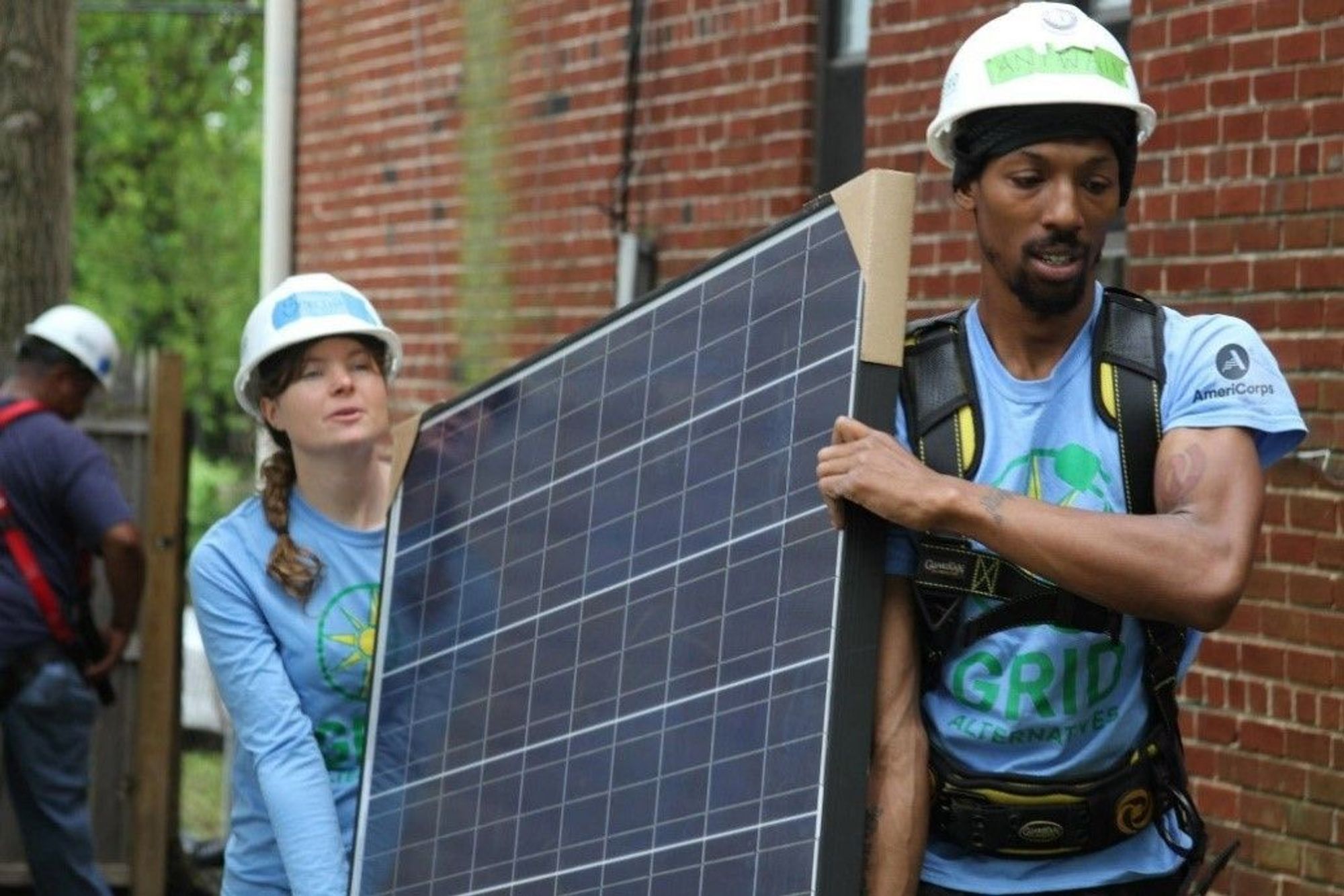 Two volunteers in blue shirts and white hard hats carry a solar panel.