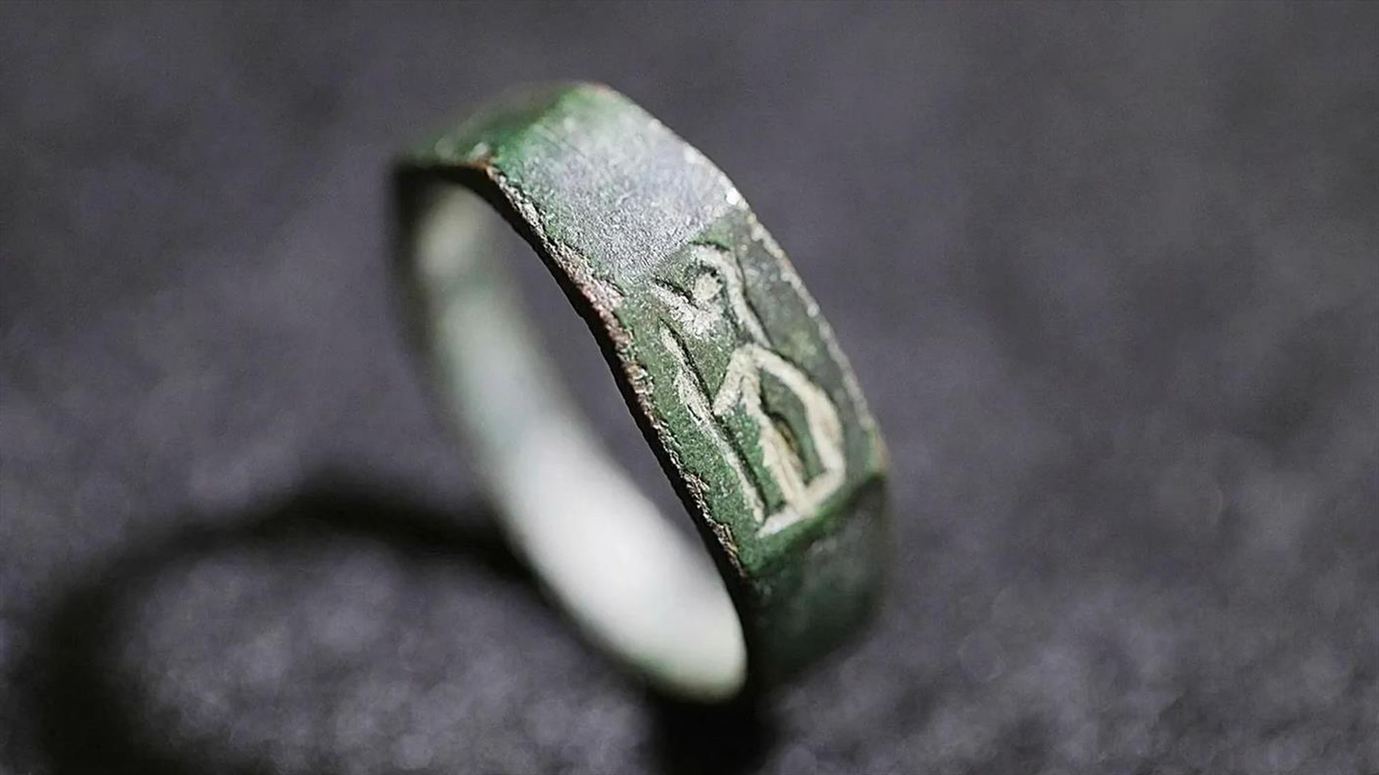 Macro color photograph: ancient ring on a muted surface. It looks especially green from oxidation in this picture. It is positioned standing upright, rather than lying flat, in 3/4 profile with the crudely etched figure of Athena/Minerva oriented toward the viewer.