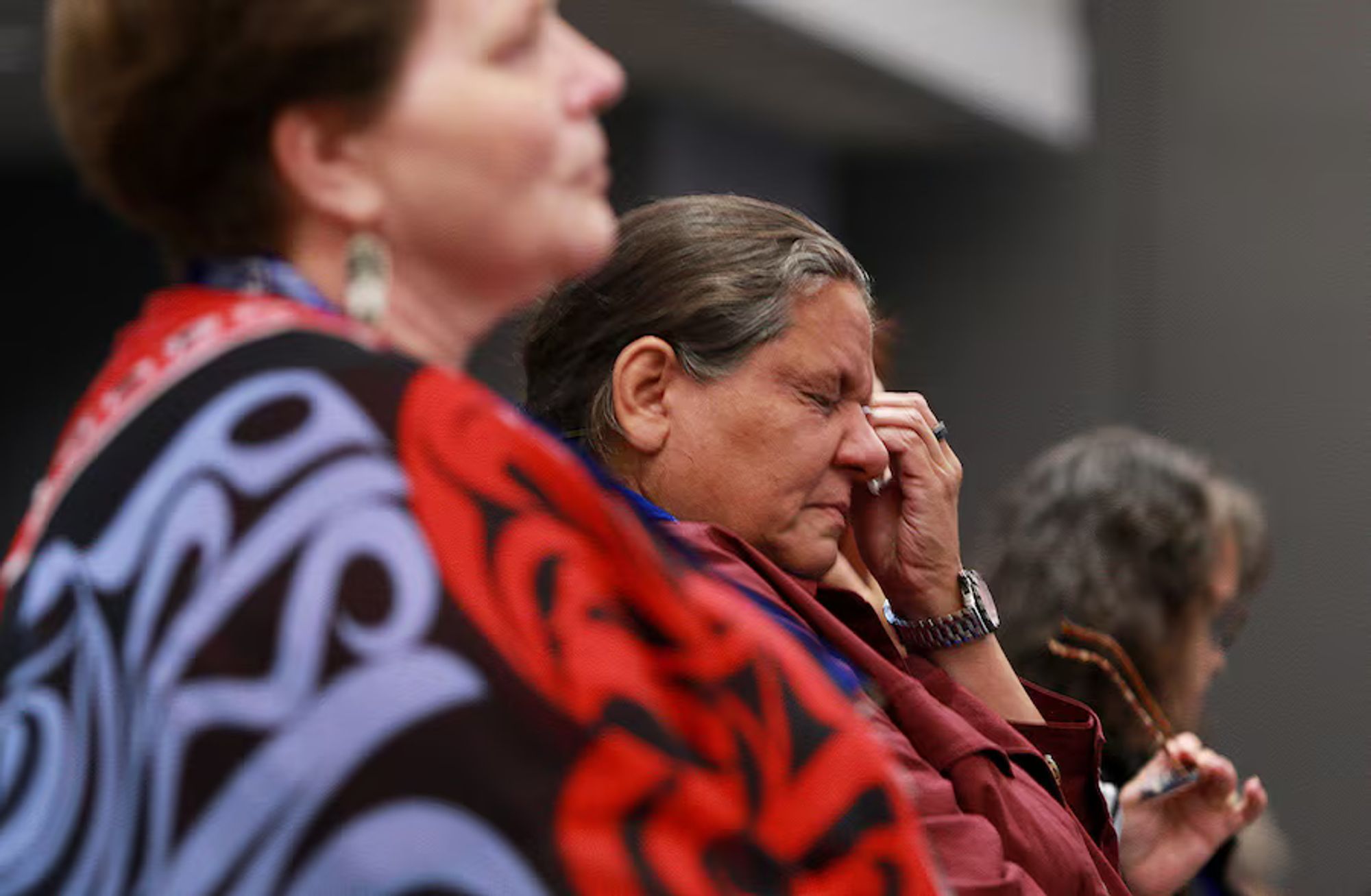 Color photo: an Indigenous woman, partly obscured by another woman (out of focus) standing next to her, wipes her shut eyes with a crumpled tissue.

Source caption reads: "Alex Wilson from the Opaskwayak Cree Nation is brought to tears during a presentation while attending the Canadian Medical Association's ceremony at the Victoria Conference Centre in B.C., Sept. 18 to formally apologize for harms to Indigenous Peoples.
Chad Hipolito/The Globe and Mail"