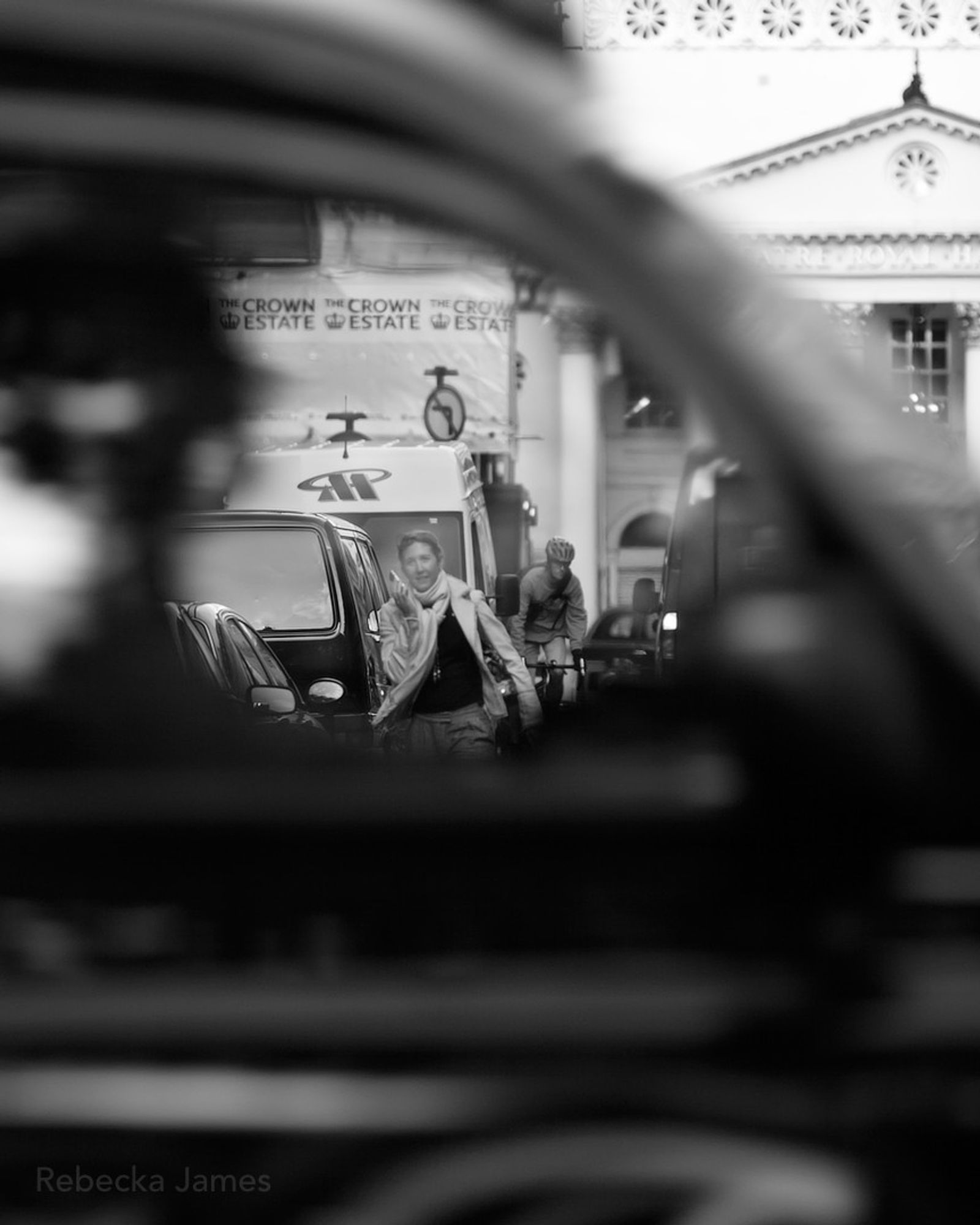 Seen through the window of a passing taxi, a woman talking on her phone walks towards the camera along Charles II Street.  Behind her is a cyclist, while the edge of a red of a Royal Mail van can be seen to their right heading the opposite way towards the Theatre Royal Haymarket.