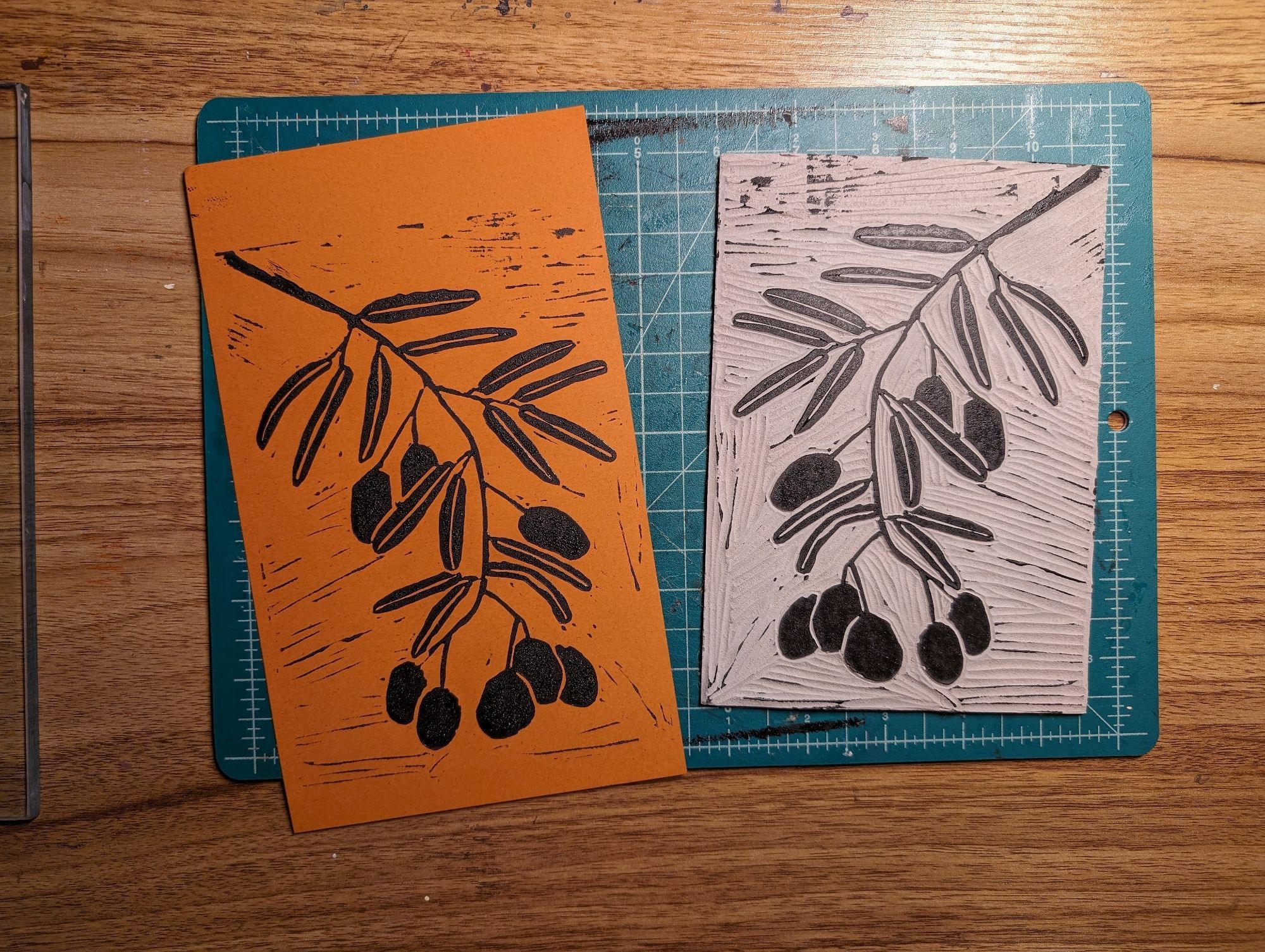 Side by side, a lino print on brown paper, and the lino pad it was carved from. The art is in black ink and shows a branch of an olive tree. The art and pad are resting on a lines carving mat, which is on a wood patterned table.