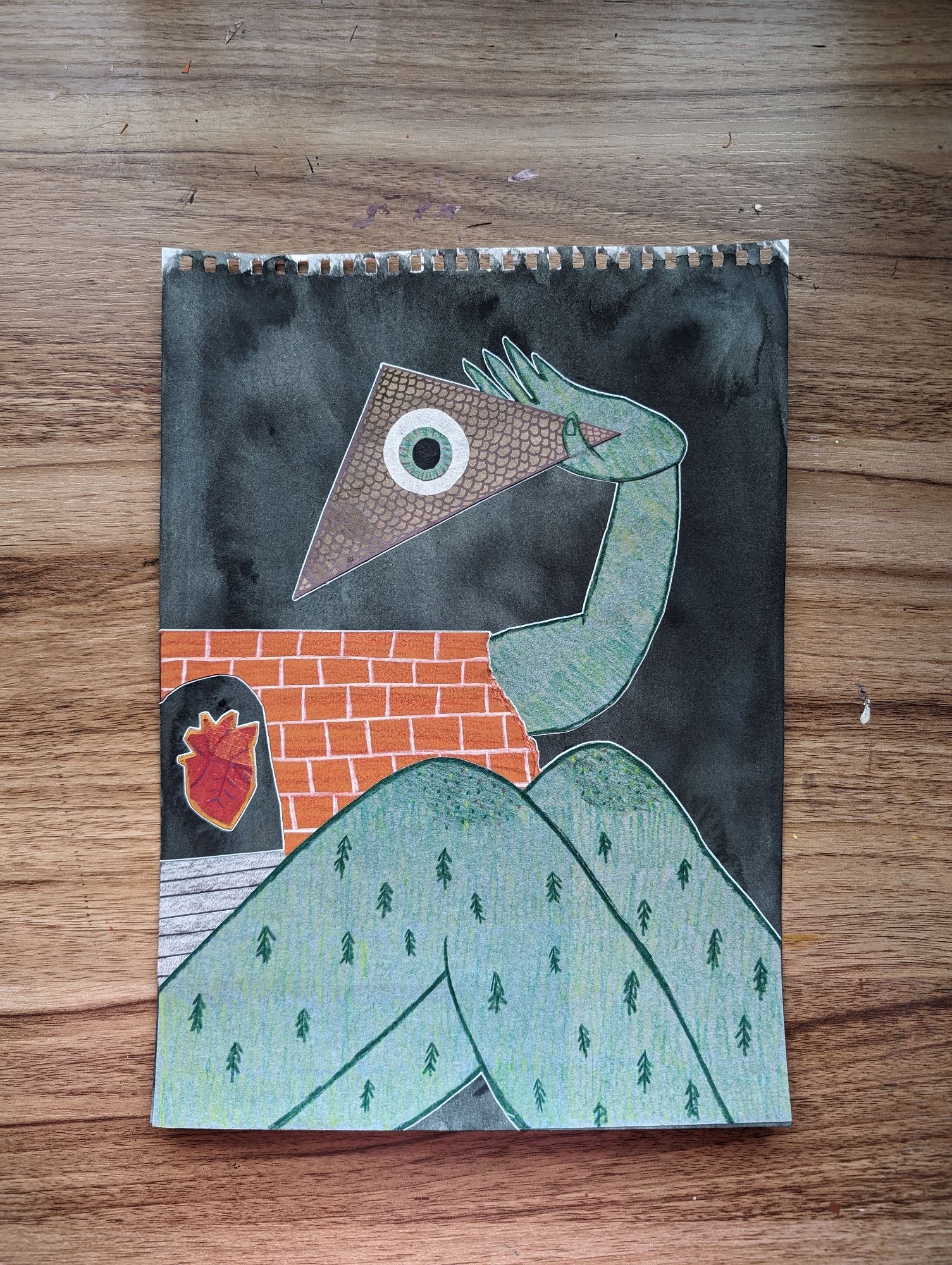 Mixed Media artwork resting on a wood patterned table. The artwork is of what appears to be a sentient building - the torso made of brick with a rounded threshold showing an anatomical heart. The large legs at the bottom are green, with small trees in place of leg hair. There is an arm holding up the brown roof of the house, and in the center of the roof is a singular eyeball with a green iris.