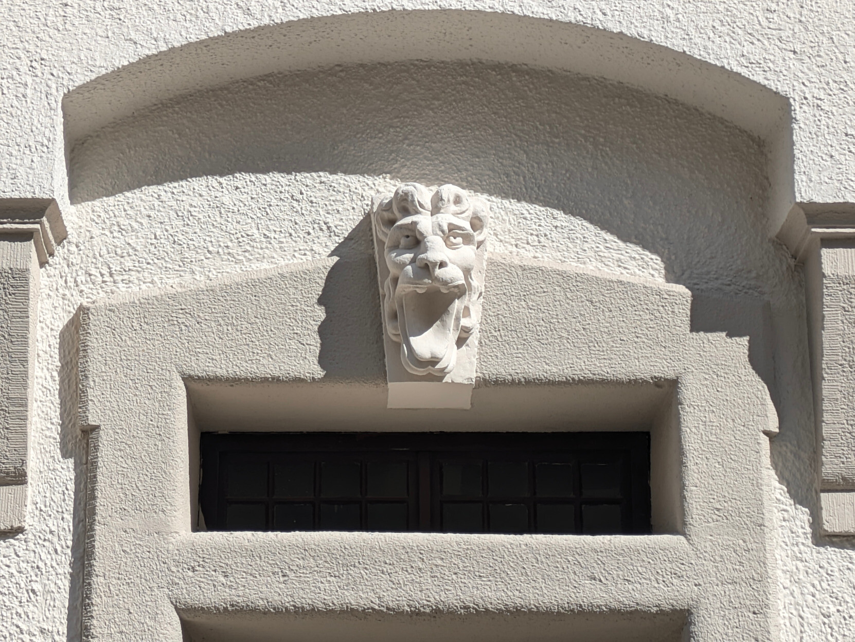 Farbfoto Querformat in weißen und grauen bis schwarzen Tönen eines Hauseingangs über der Tür: In einer oben gebogenen Nische über dem verputzten Rahmen um Oberlicht und Tür hängt eine weiße Skulptur vom Kopf eines leicht stilisierten brüllenden Löwen mit offenem Maul. Das Sonnenlicht kommt von rechts oben und sorgt für die Schlagschatten von Nische und Figur.