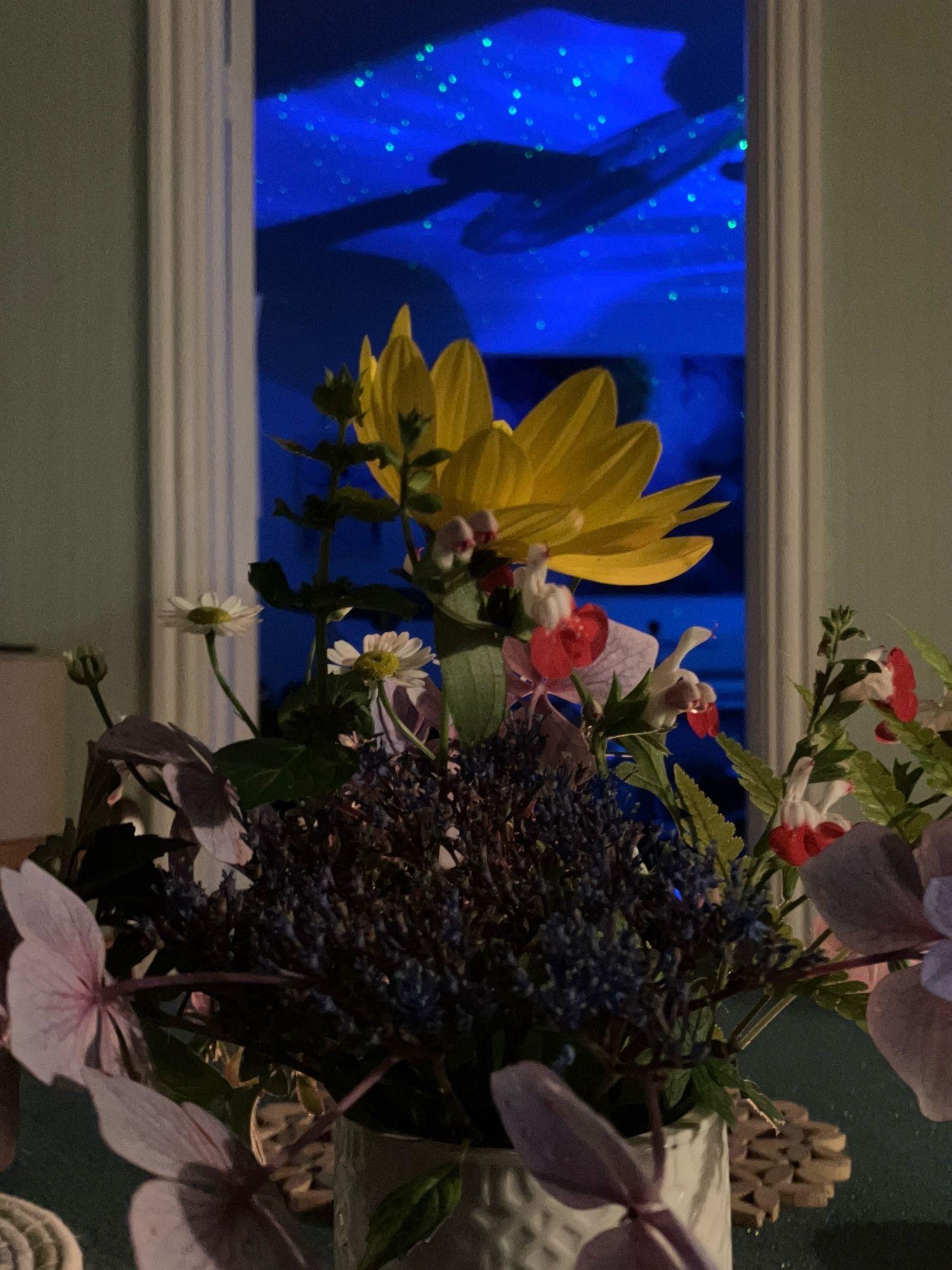 A cylindrical vase with flowers from the garden sits on a table. A yellow flower is looking up from the bunch. In the background, through the doorway, the room is bathed in blue light