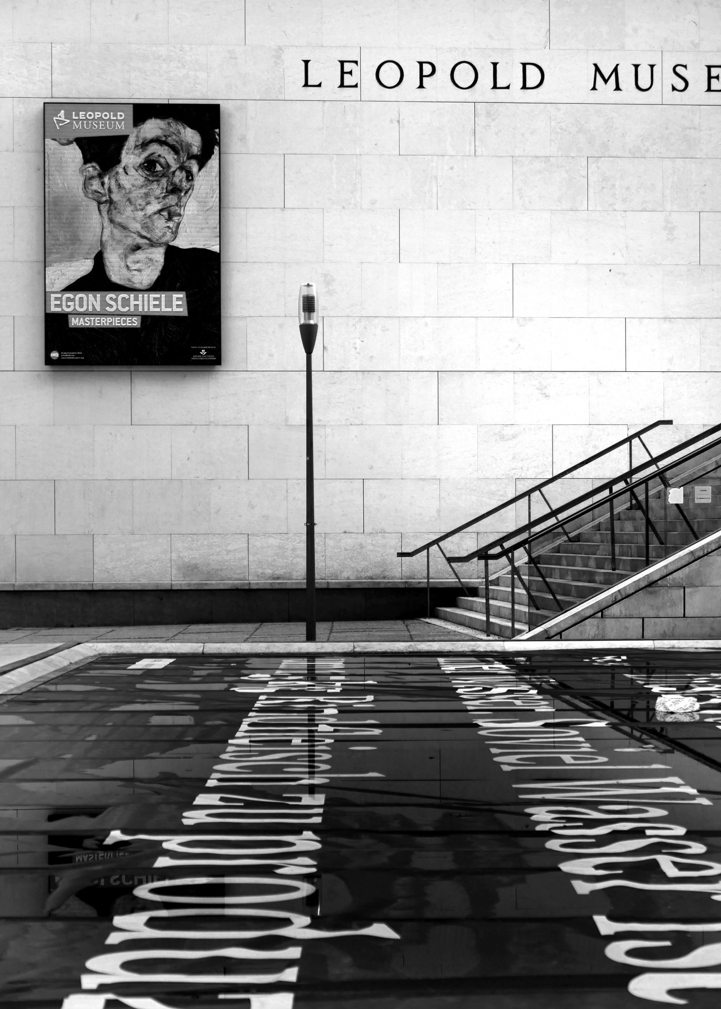 Aufgang zum Leopoldmuseum, Wien. Im Vordergrund ist ein gemauerter Teich, abgedeckt mit einer schwarzen Plane mit weißer, großer Schrift. An der Museumswand hängt ein Plakat von der Egon Schiele Ausstellung. Rechts im Hintergrund, neben einer Laterne, ist die Treppe zum Museumseingang.