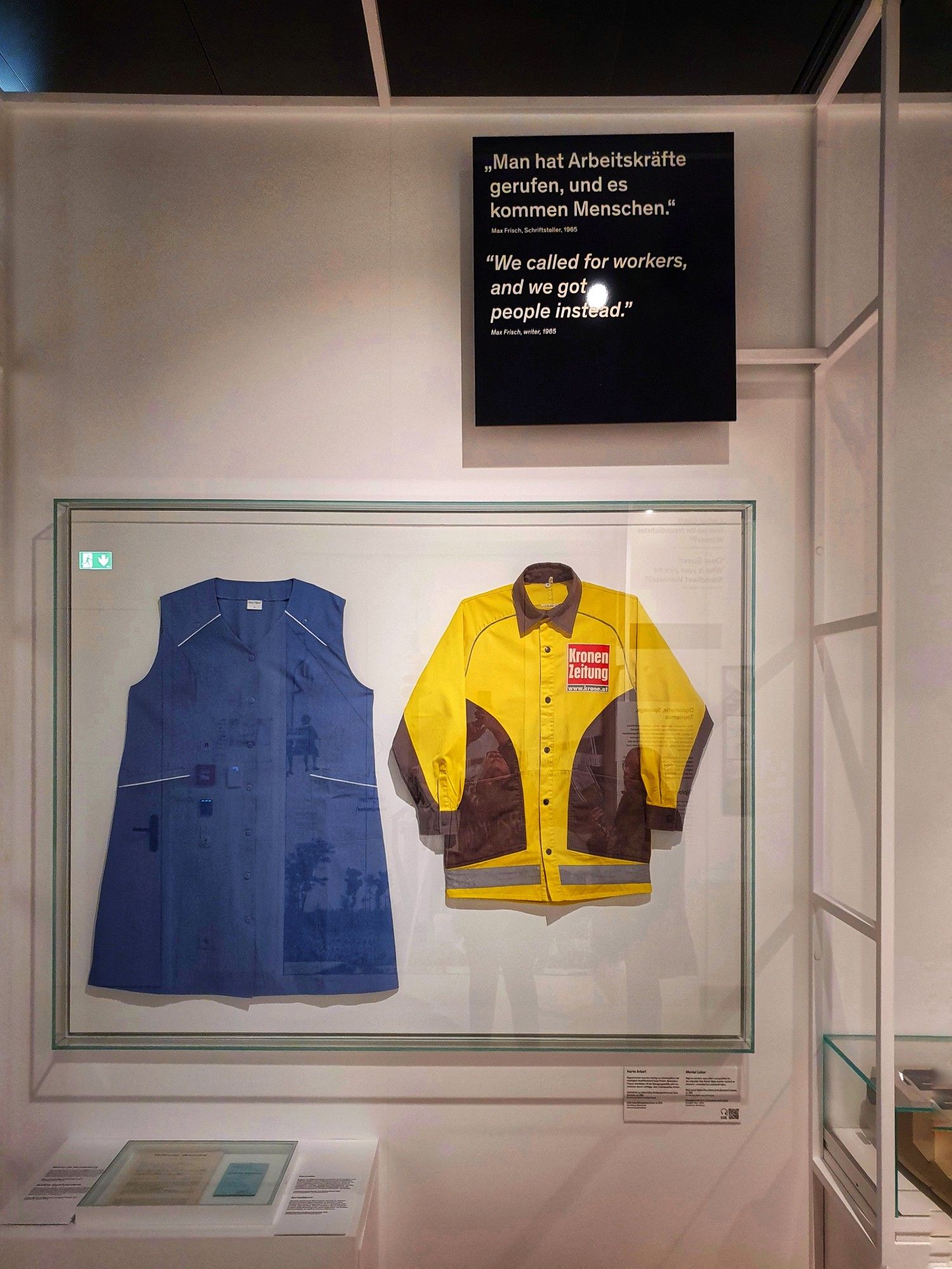 In einer Vitrine hängen der Arbeitskittel einer Reinigungsfrau und die Regenjacke eines Zeitungsverkäufers.
Auf einer Tafel steht eine Zitat: 'Es wurden Arbeitskräfte gerufen, und es kommen Menschen'.