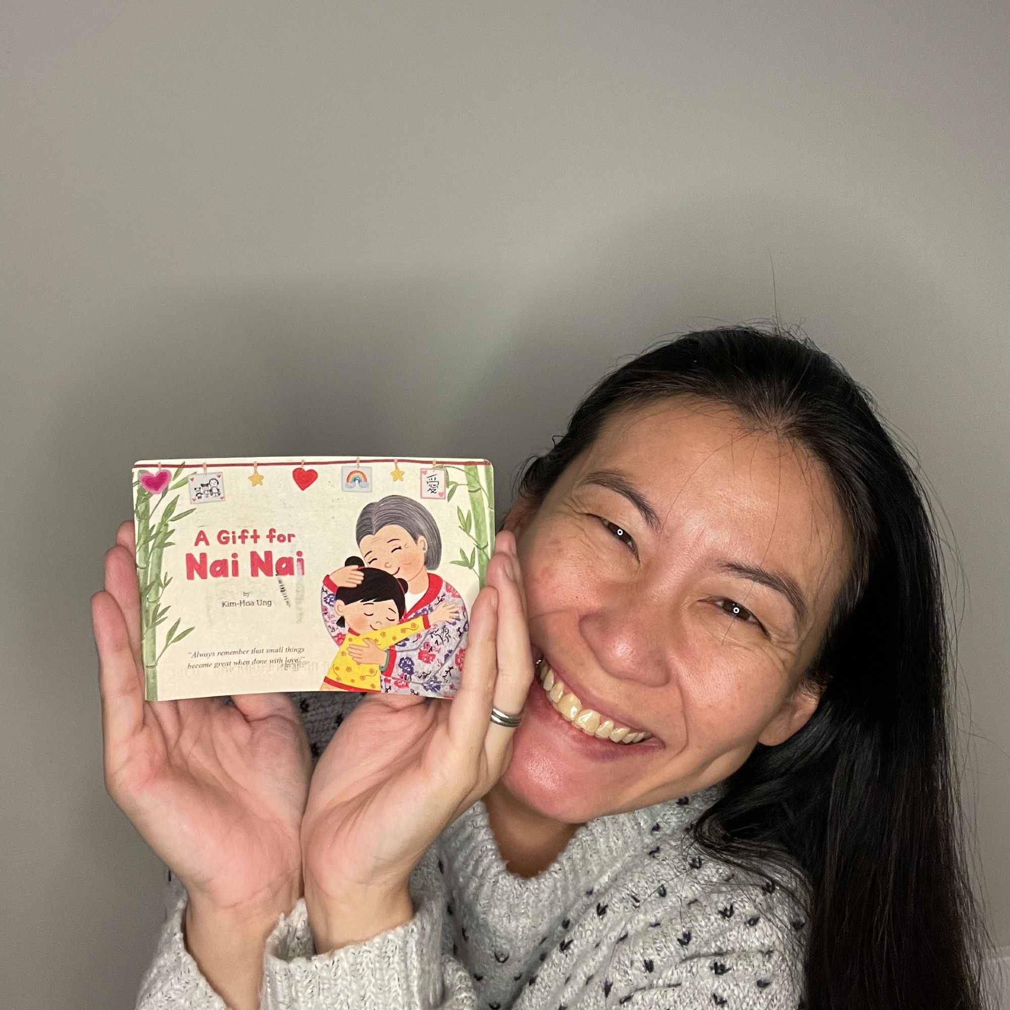 Picture of Sylvia Chen holding up a postcard from Kim-Hoa Ung. The postcard has an illustration of Kim-Hoa's upcoming debut picture book A GIFT FOR NAI NAI, with a little girl with black hair pulled up into a cute two-bun hairstyle wearing a yellow shirt trimmed with red, hugging her grandma Nai Nai.