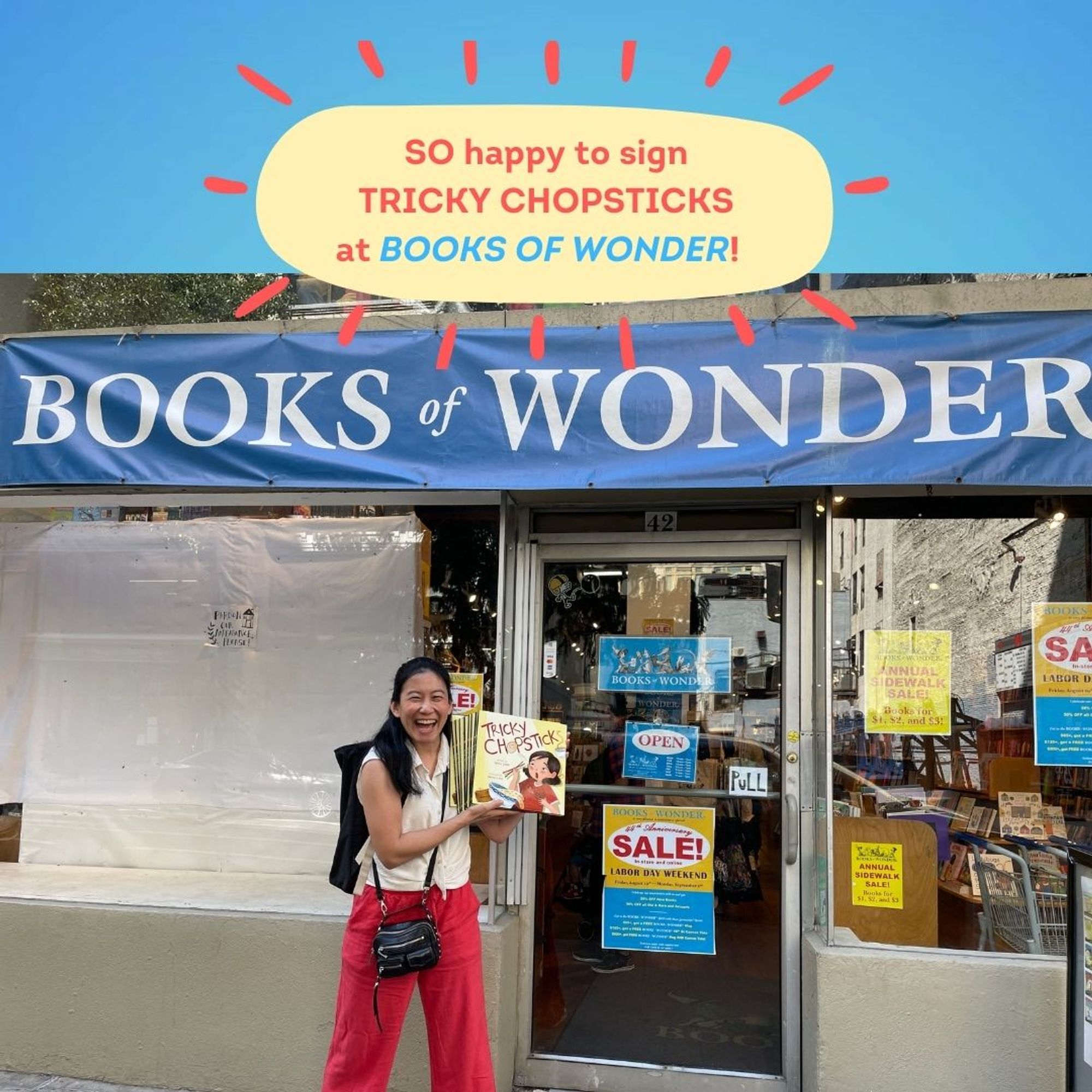 Picture of author Sylvia Chen in a white top and red pants holding up signed copies of TRICKY CHOPSTICKS in front of Books of Wonder in NYC.