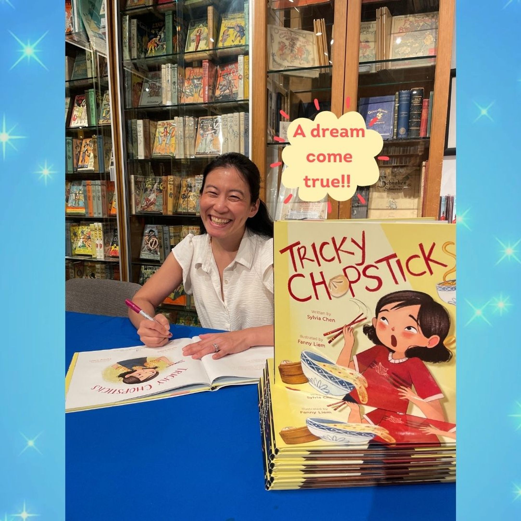 Picture of author Sylvia Chen signing copies of TRICKY CHOPSTICKS on a blue tablecloth inside Books of Wonder, with a light yellow cloud callout with red dashes flaring out saying "A dream come true!!"