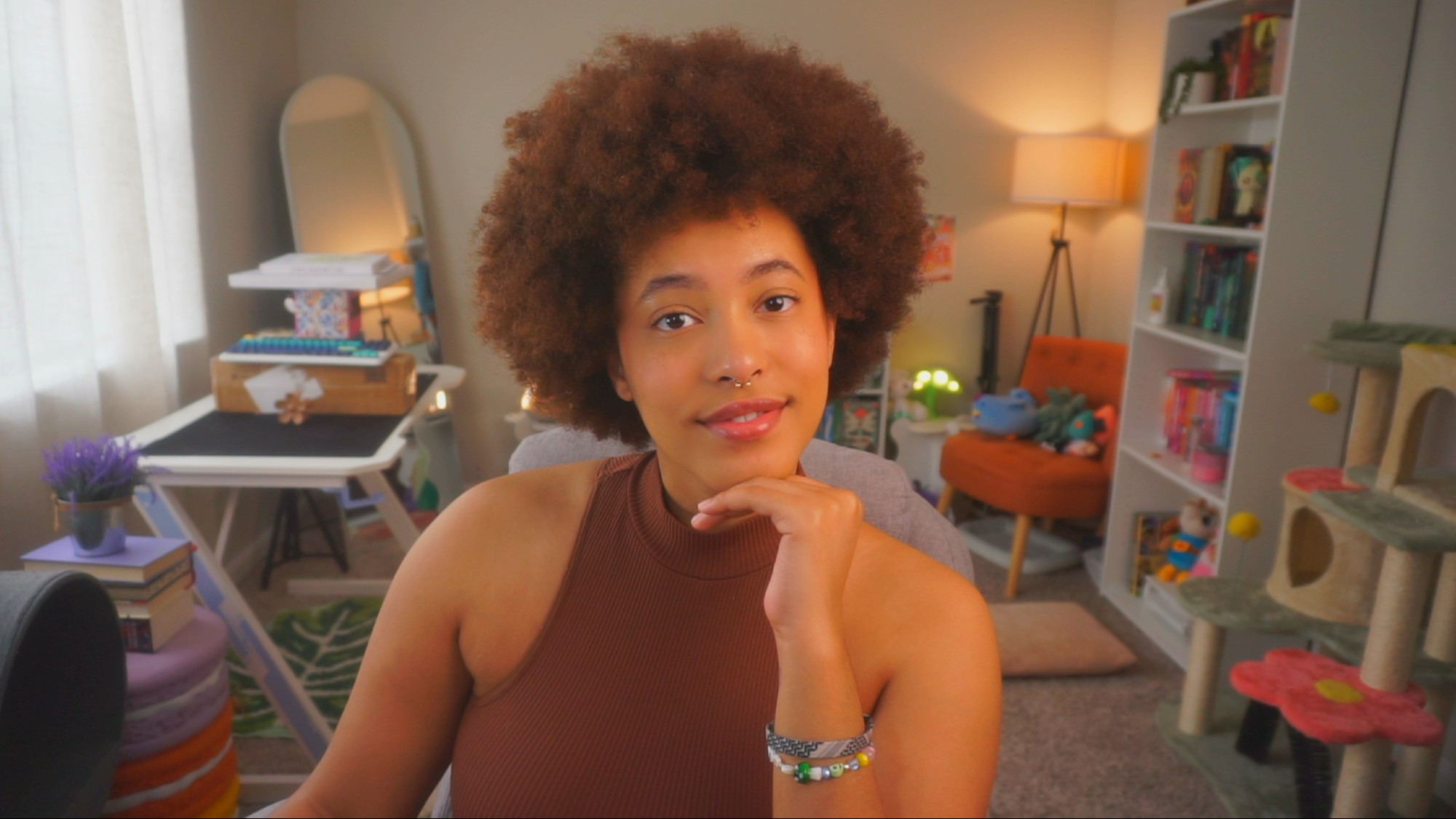 Sage sitting at their desk while wearing a brown sleeveless shirt. Sage's hair is in a large afro and their glossed lips are slightly parted.