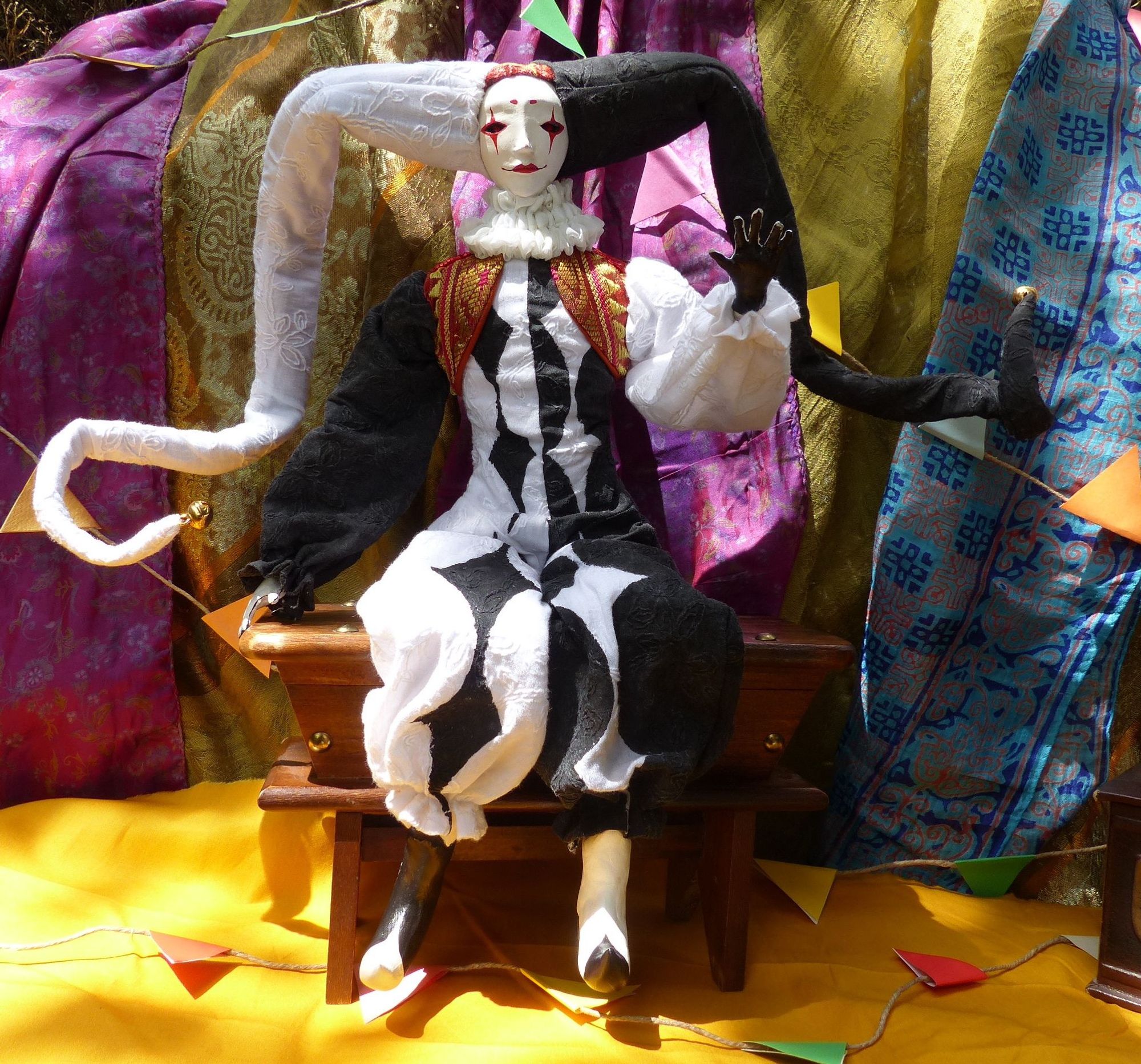 Full body picture of a handcrafted posable soft jester doll with an asymetrical black and white diamond overalls costume and a red and gold vest. The doll is sitting on a little bench and waving