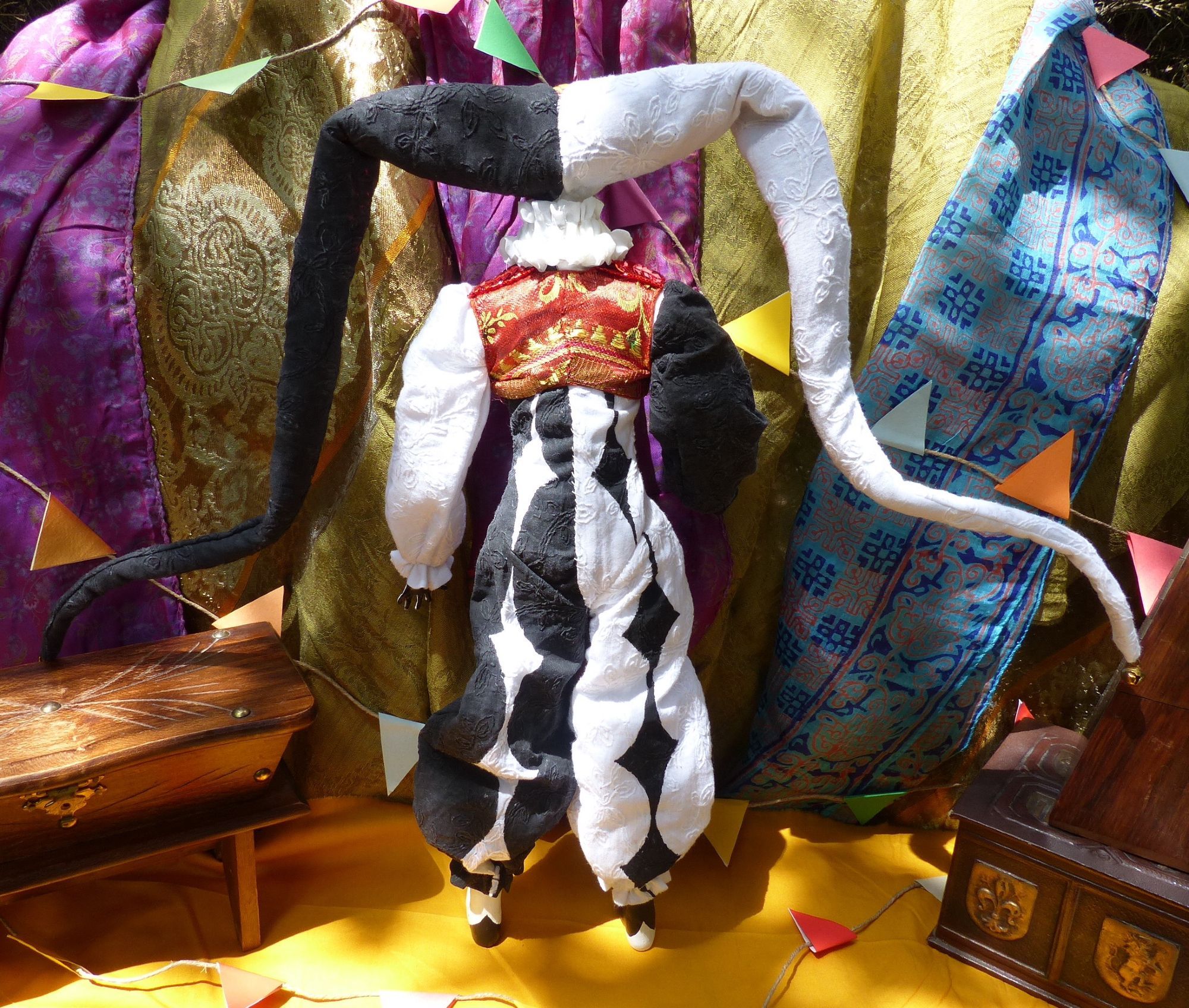 Full body picture of a handcrafted posable soft jester doll with an asymetrical black and white diamond overalls costume and a red and gold vest. The doll is facing backward.