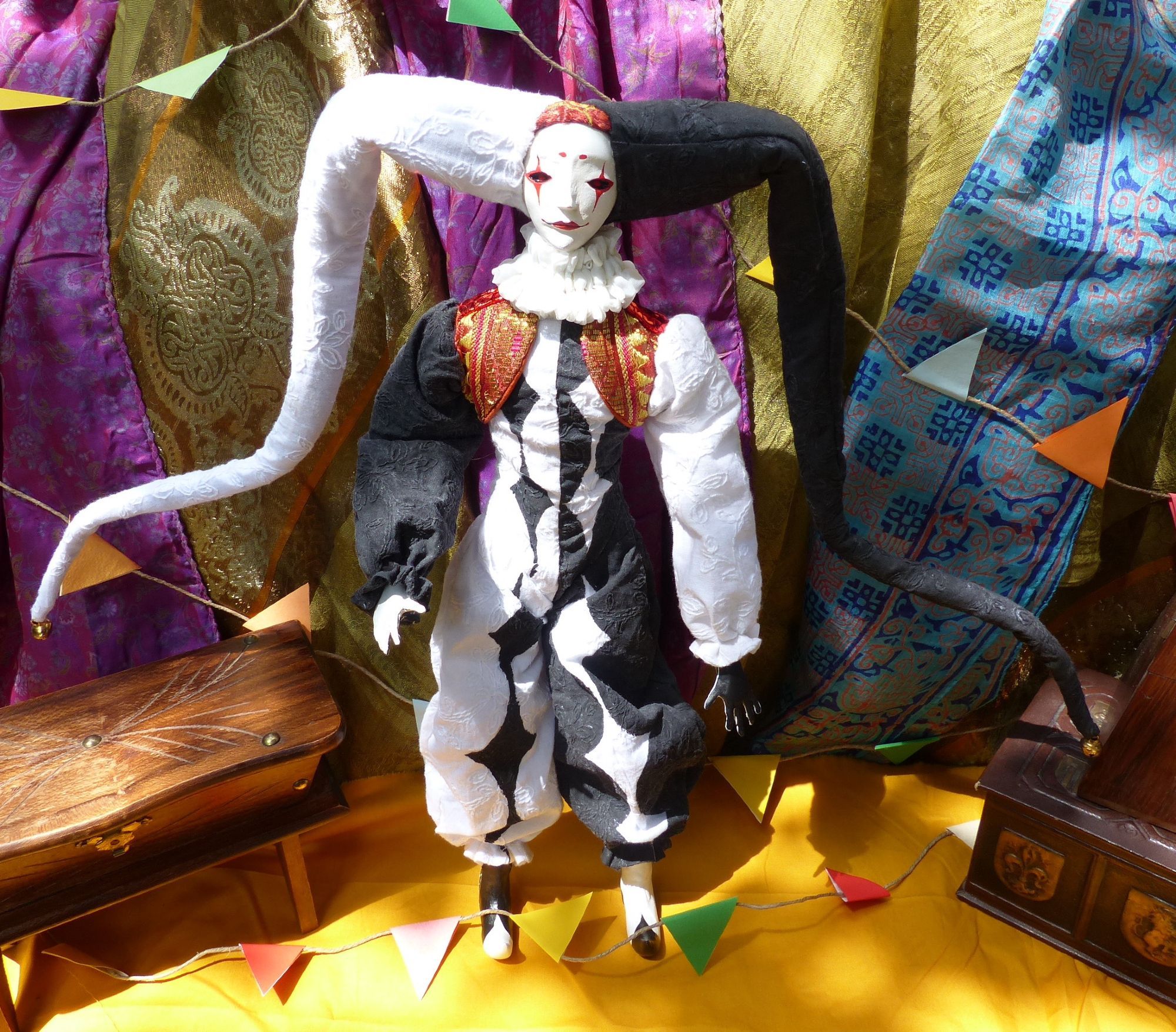 Full body picture of a handcrafted posable soft jester doll with an asymetrical black and white diamond overalls costume and a red and gold vest. The doll is facing forward.