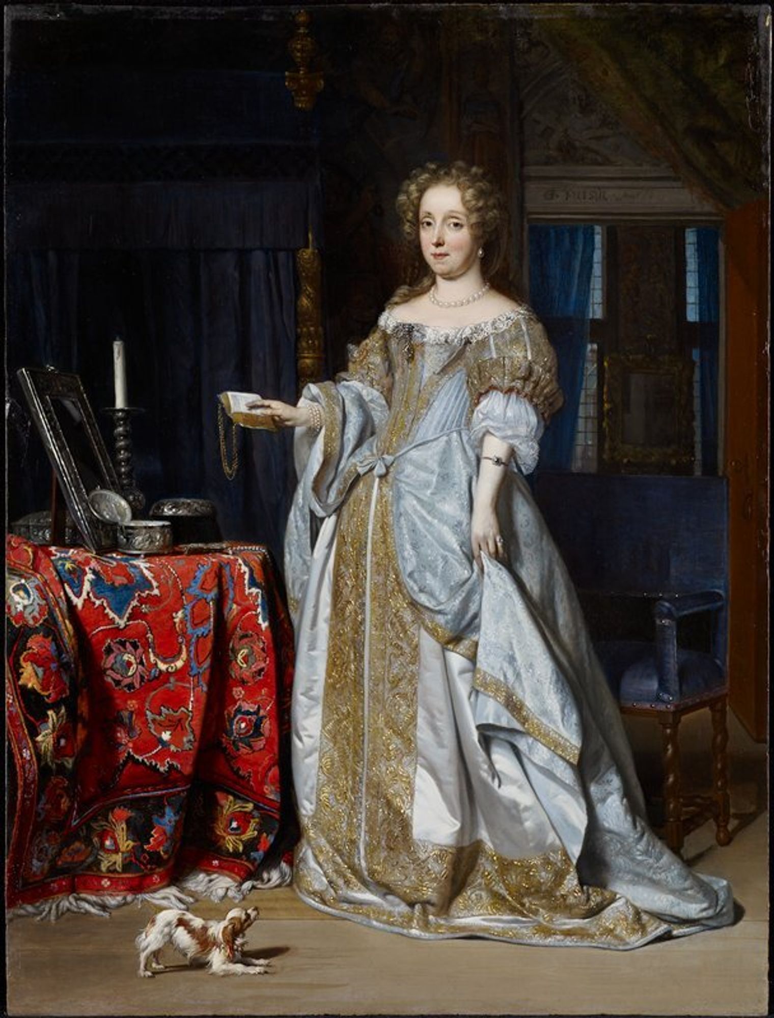 Portrait of a woman. Dutch golden age. A lady about thirty-four years of age with her fair hair in curls, stands full length at her toilet table, almost facing the spectator. She has a book in one hand and holds up her skirt with the other. She wears a light blue satin dress embroidered with gold lace. On the table, which is covered with a Turkey carpet, are a mirror, a silver box, and a candlestick. A spaniel frisks at her feet.
We can now put a name to this face. For centuries, this painting was known simply as Portrait of a Woman. Recent research revealed her identity: Lucia Wijbrants, the wealthy widow of a Dutch cloth merchant. She stands at her dressing table, surrounded by her pricey possessions, while a frisky spaniel begs for attention. Lucia emerged from a variety of sources, including known portraits (of her and her husband) featuring similar likenesses and some of the same belongings pictured here. In fact, the bed behind her was previously owned by her husband’s first wife.