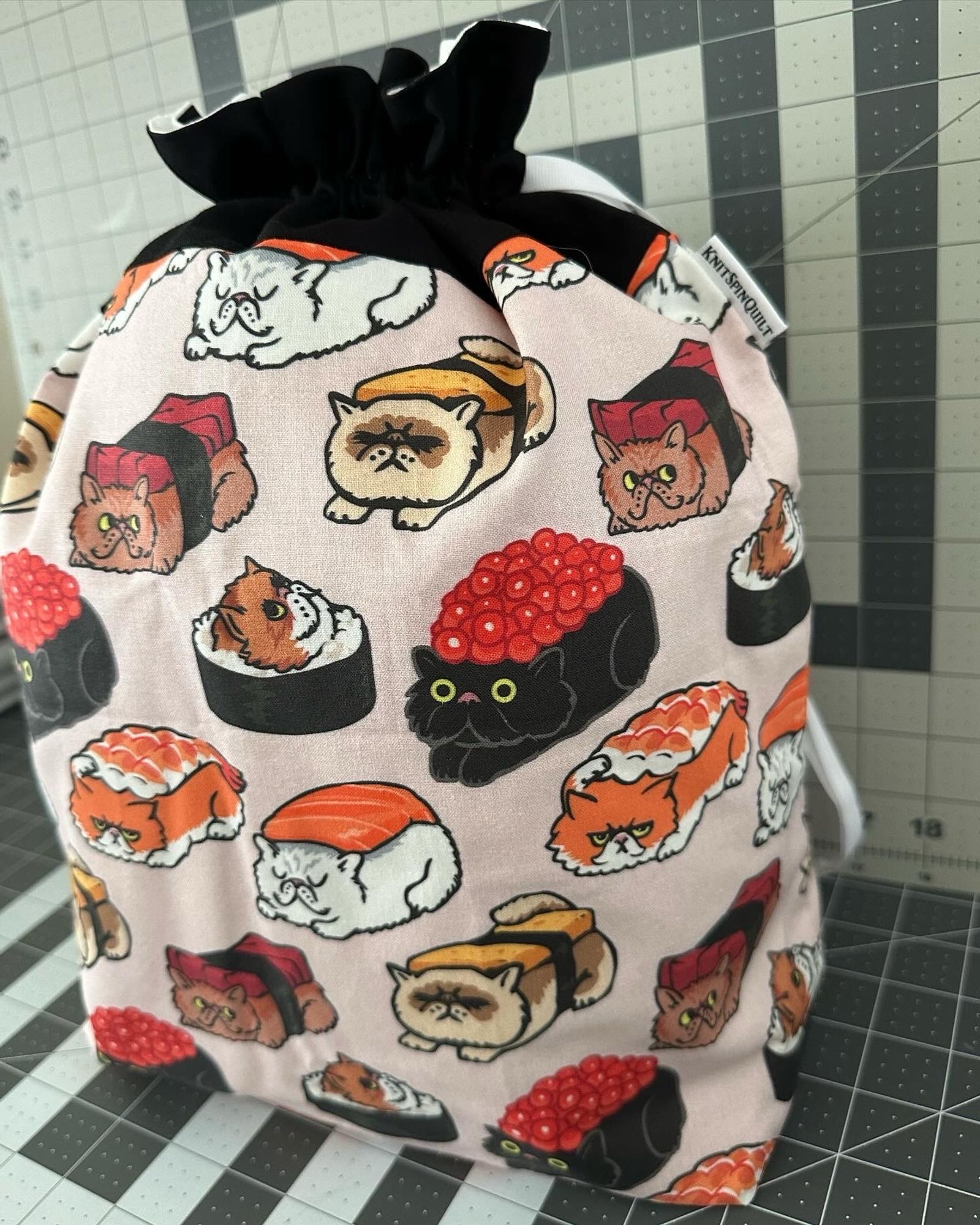 A large drawstring bag standing up on a gridded cutting mat. The top is black, and the body of the bag is pale pink with line art of various kinds of cats dressed up as sashimi