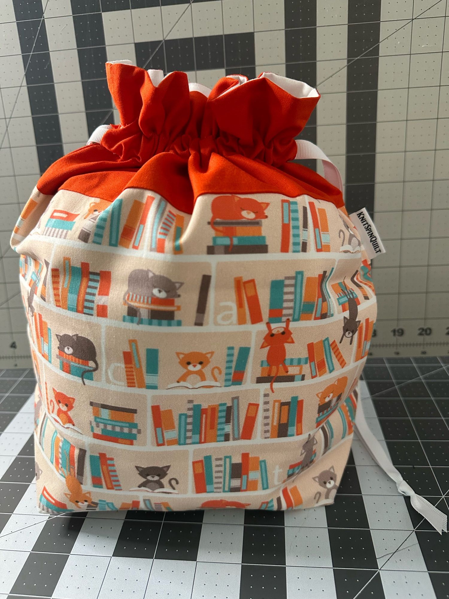 A medium drawstring bag with a solid orange top standing stuffed on a gridded cutting mat. The body of the bag is a pale orange with bookshelves showing cats sleeping on books