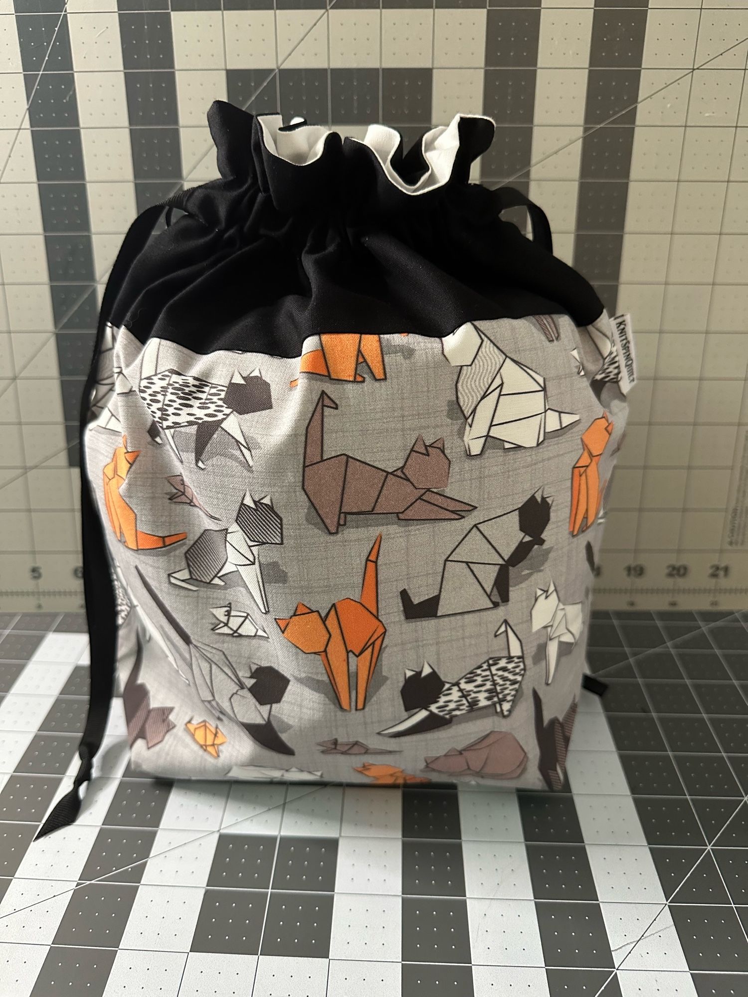 A medium drawstring bag standing stuffed on top of a gridded cutting mat. The top is black and the body fabric of the bag is grey with line art of various cats drawn as if they are origami.