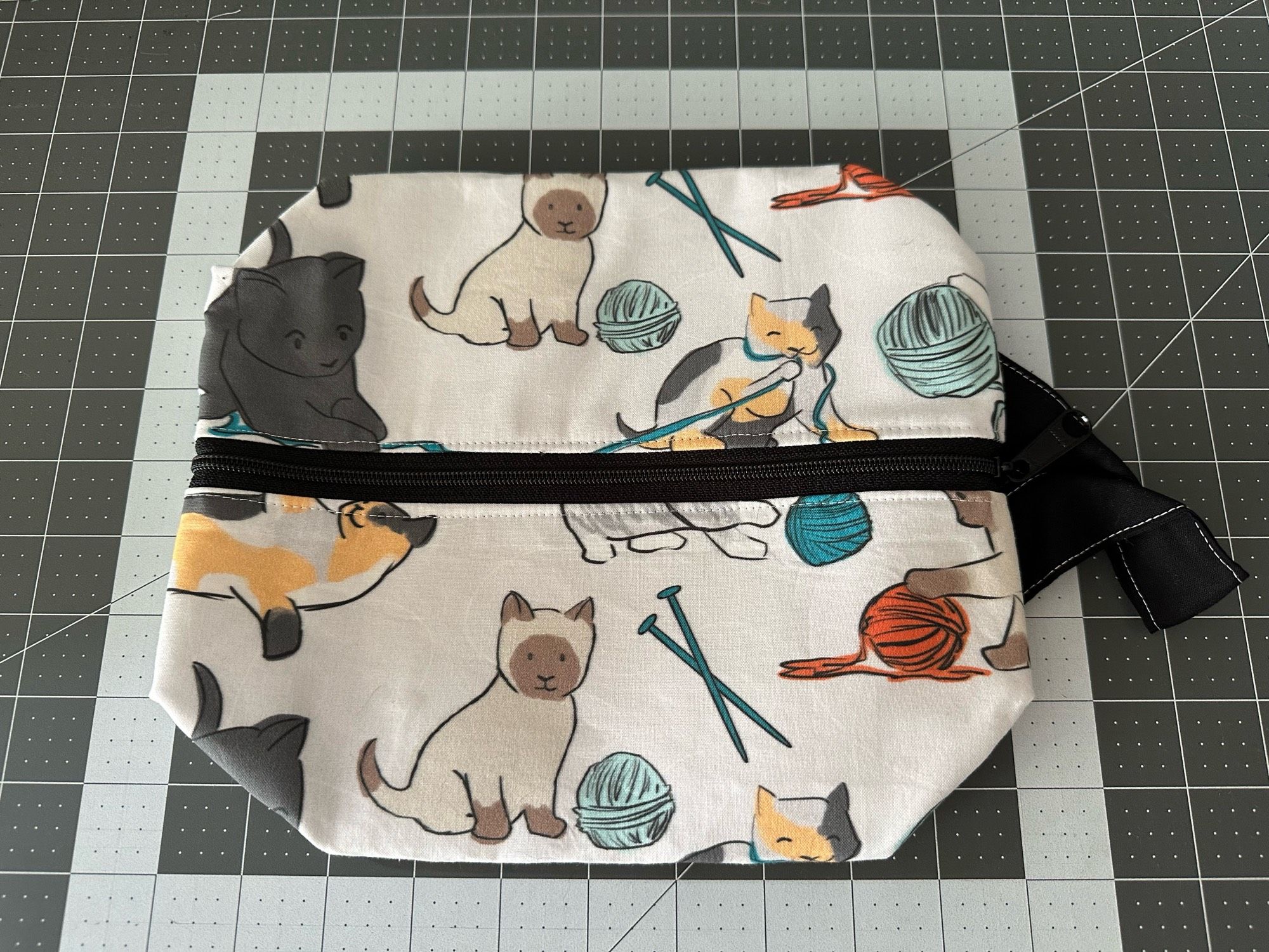 A flattened zippered box bag, white fabric with line art of cats with yarn. The zipper and handle of the bag are black.