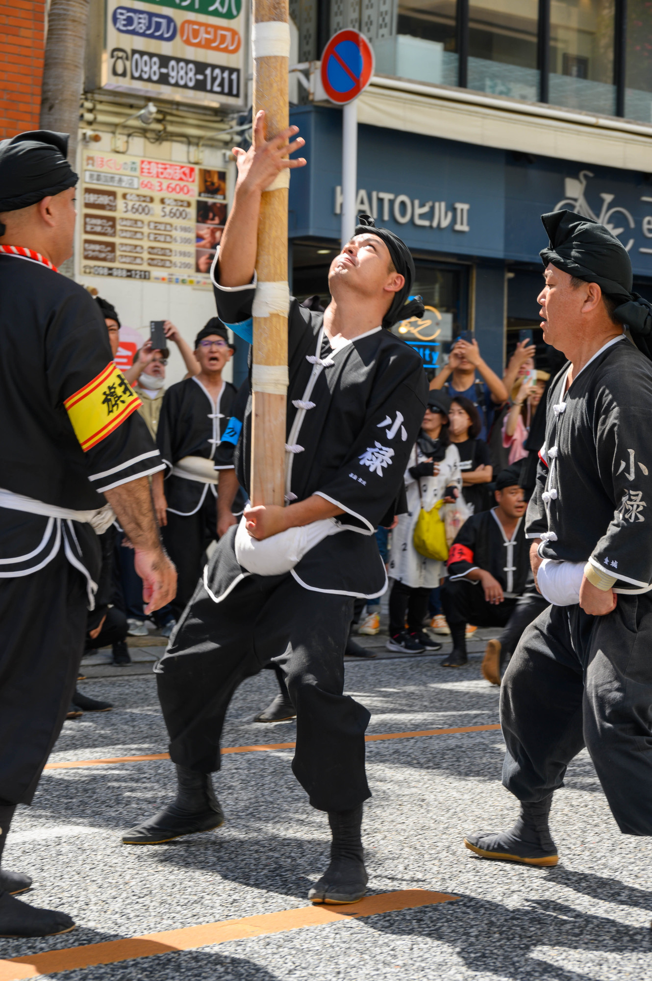 A group of men in traditional black garments participate in a festival, lifting a bamboo pole. The scene captures their effort and determination, with spectators in the background observing the event.