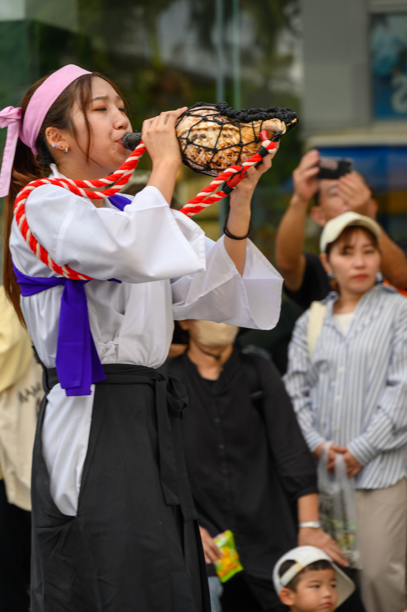 A young woman wearing traditional attire plays a large shell horn, surrounded by a small audience. Some spectators are taking photos while others watch attentively.