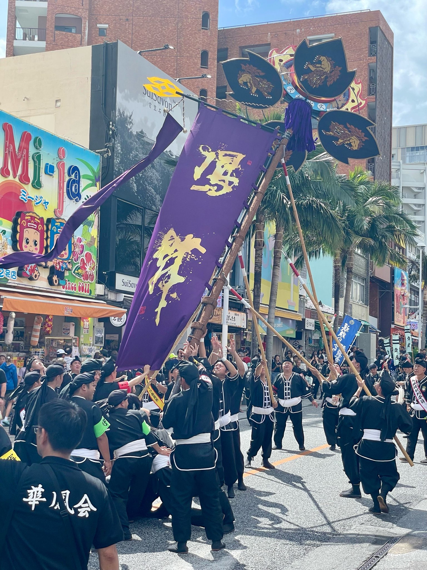 A group of people dressed in traditional black clothing are participating in a festival, holding banners and poles. A colorful street scene features shops and palm trees in the background, with skies partly cloudy. The atmosphere is festive and lively.