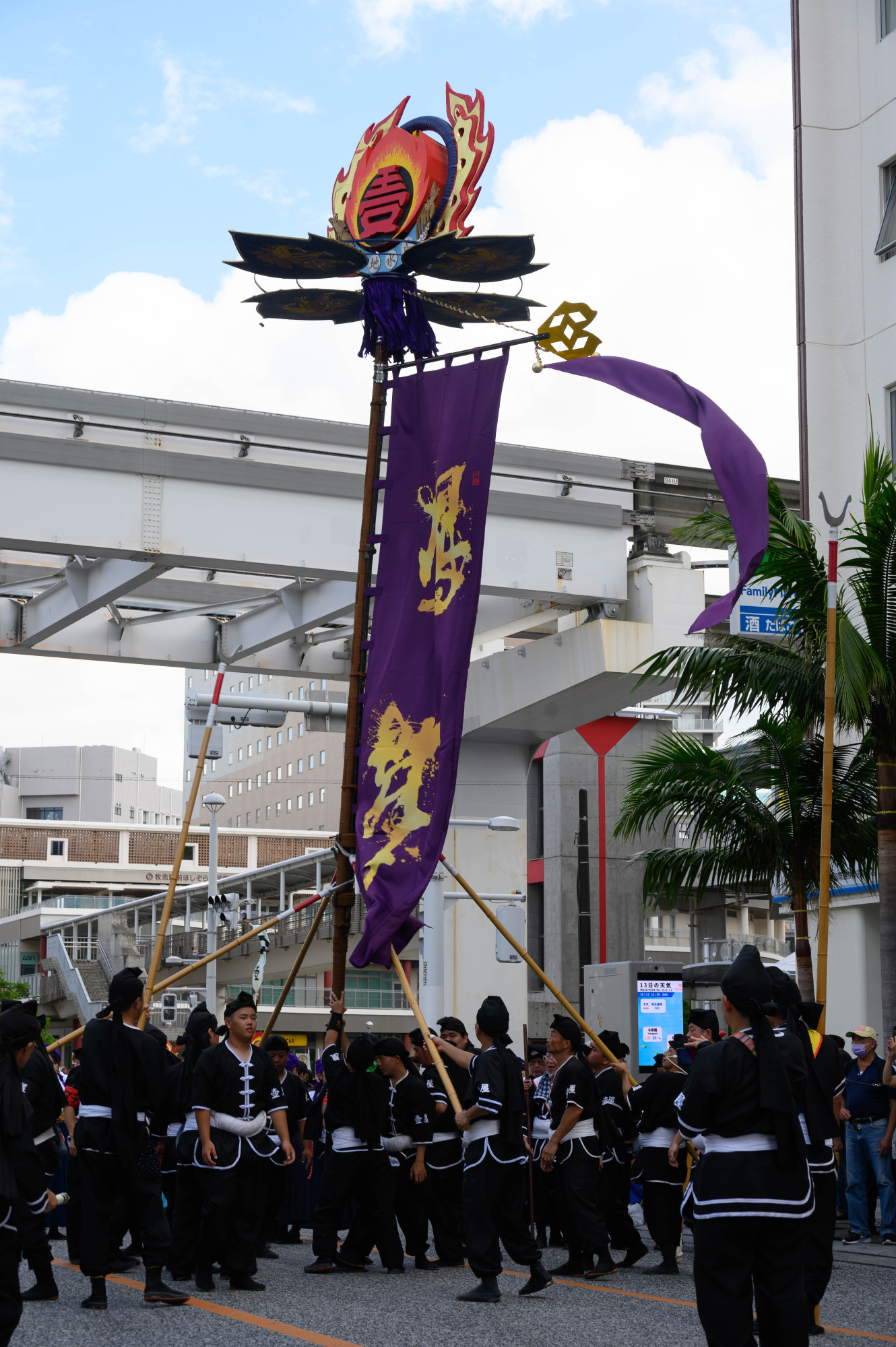 A group of people in traditional black clothing participates in a cultural event, holding a large banner and a decorated pole with flames at the top. The scene takes place outdoors, with urban buildings and a palm tree in the background.