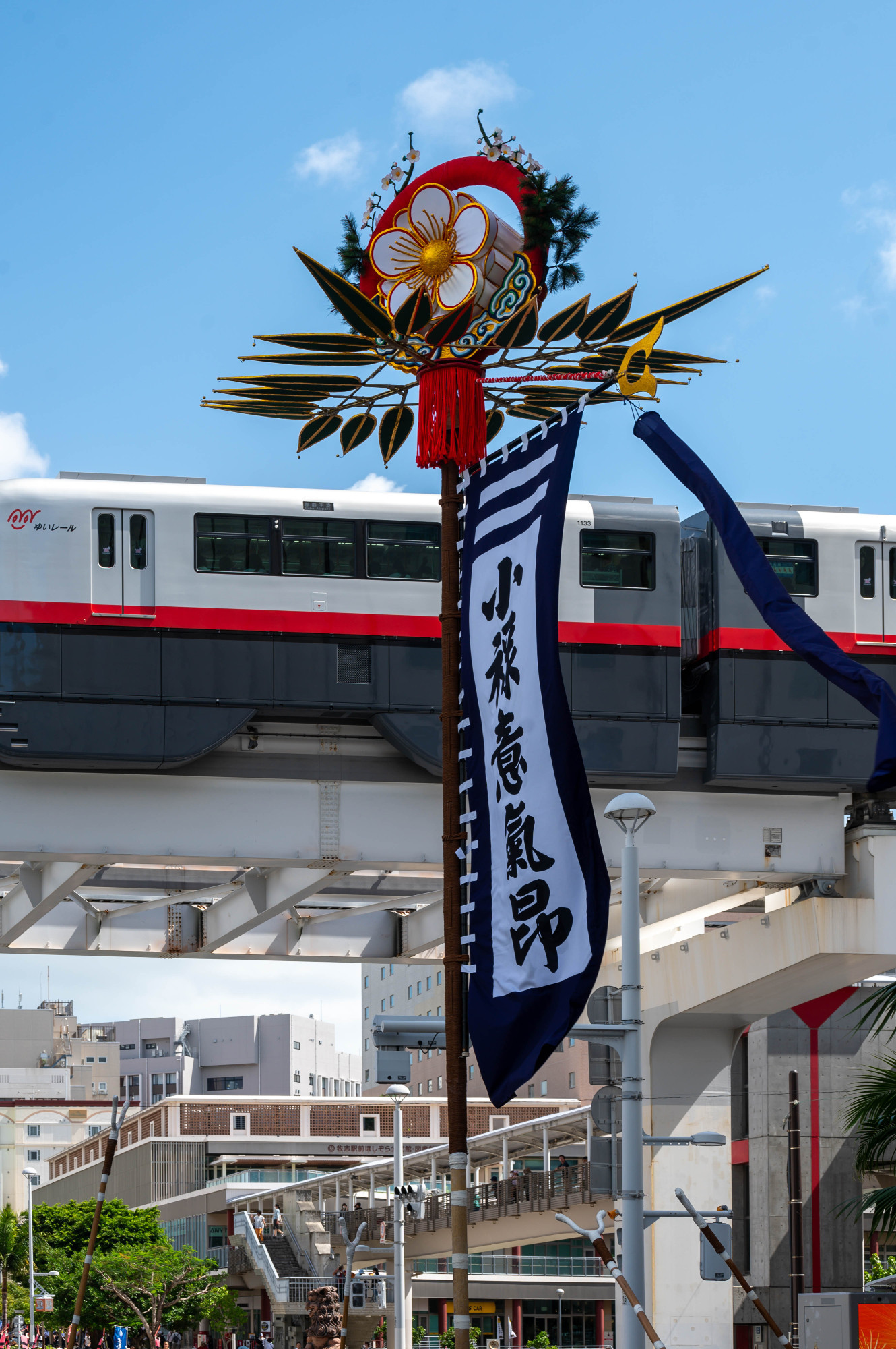 A colorful decorative pole with floral elements and a banner stands in the foreground, while a train moves along an elevated track in the background. The sky is clear with few clouds.