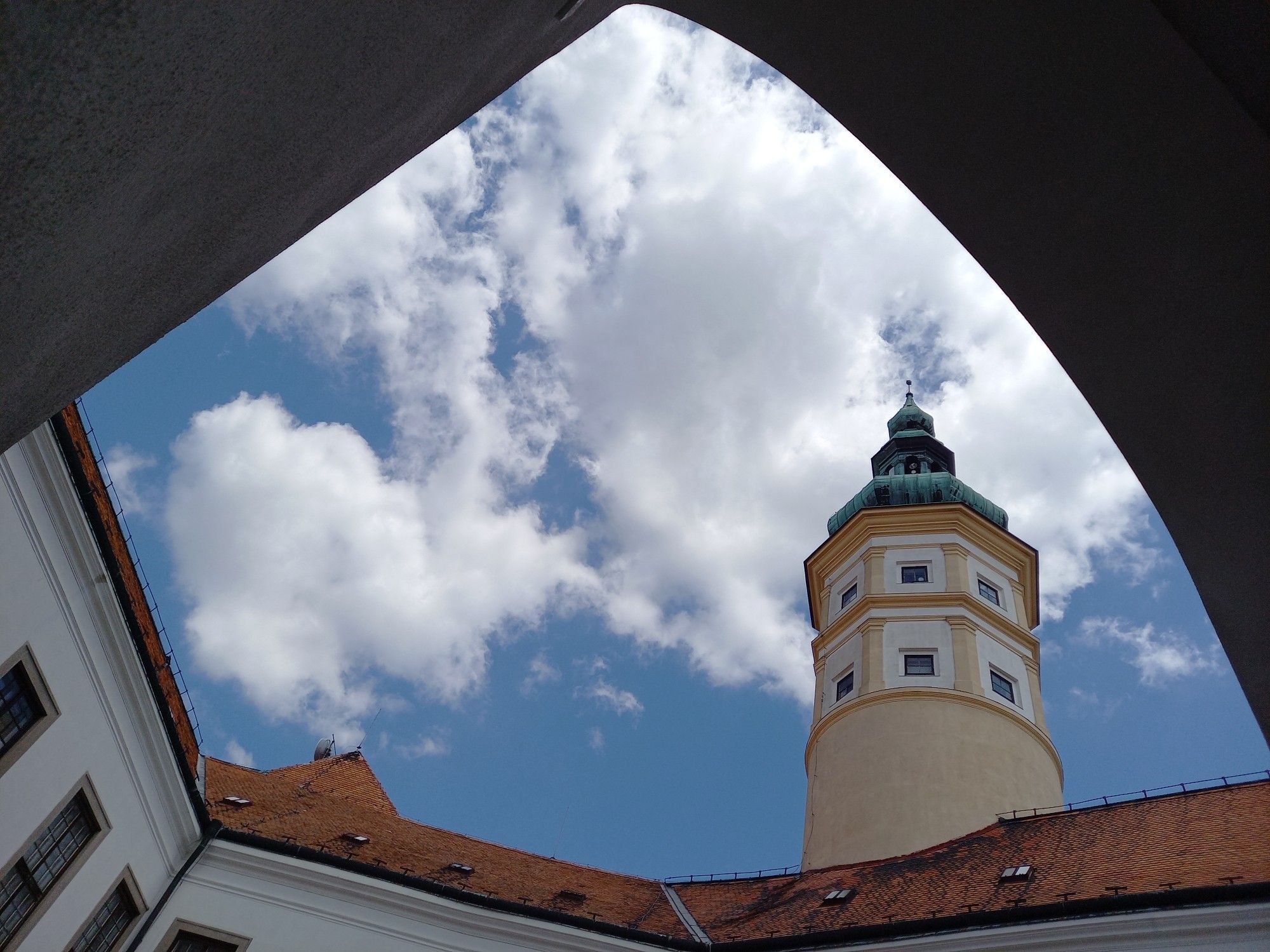 Blick aus einem Schlosshof nach oben, blau/weißer Himmel. Ziegeldächer, ein Schlossturm in weiß und Schönbrunner Gelb mit kupferner welscher Haube.