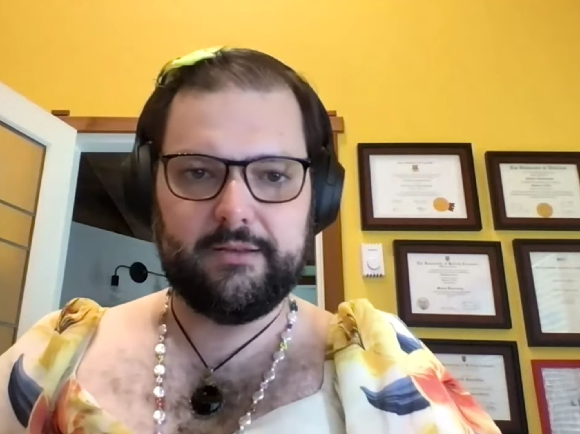 Lucía M. Polis, wearing a low-cut yellow dress with flowers, a pearl necklace, a pendant, and a yellow bow in her hair, in a yellow room, with diplomas hanging on the wall behind her