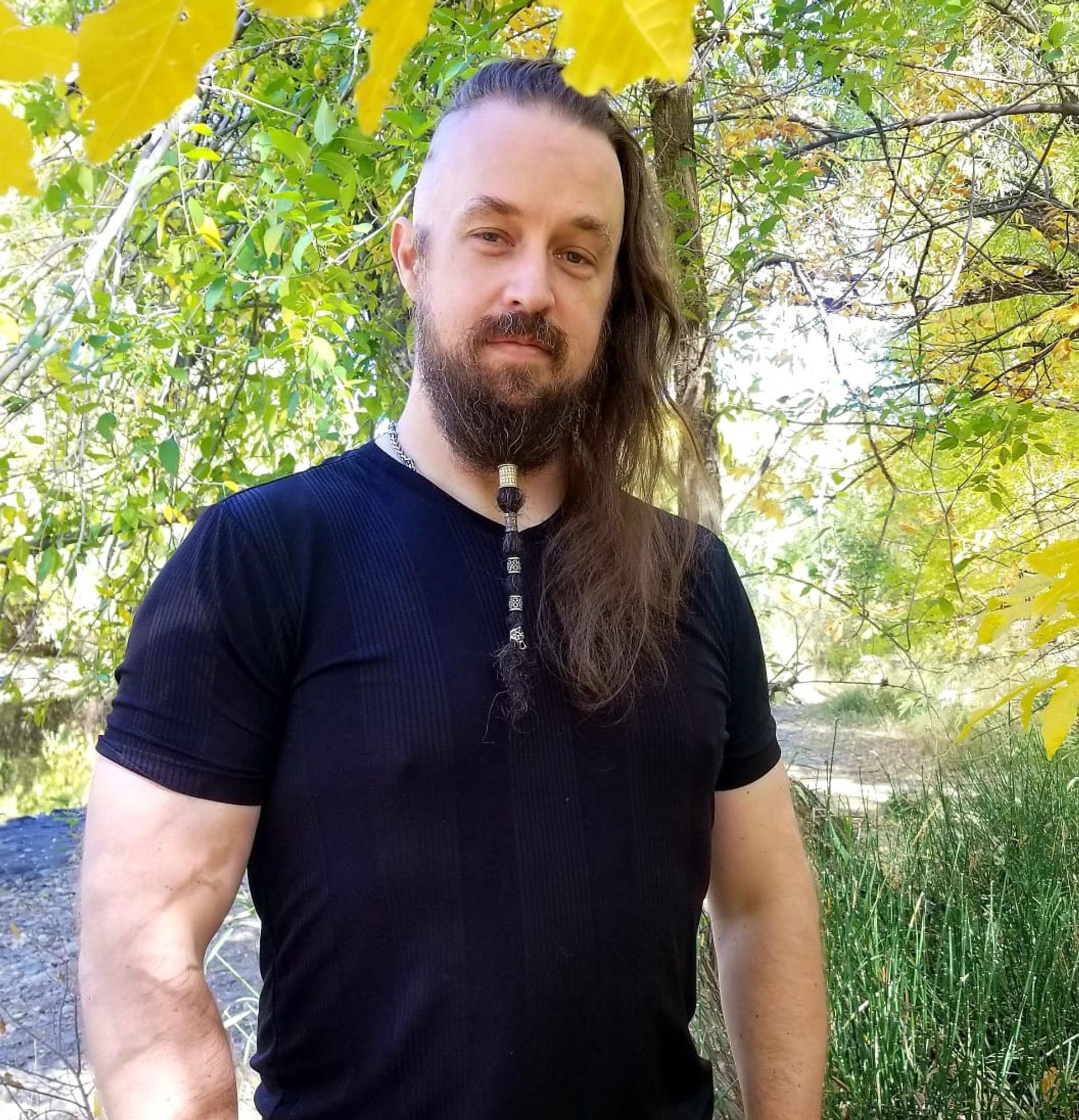 A photo of Koulnis looking warmly at the camera. He is wearing a black shirt and standing among trees with green and yellow foliage, with a stream and tall grasses in the background. He has long brown hair shaved on the sides and a chest-length beard tied with five Viking beard rings.