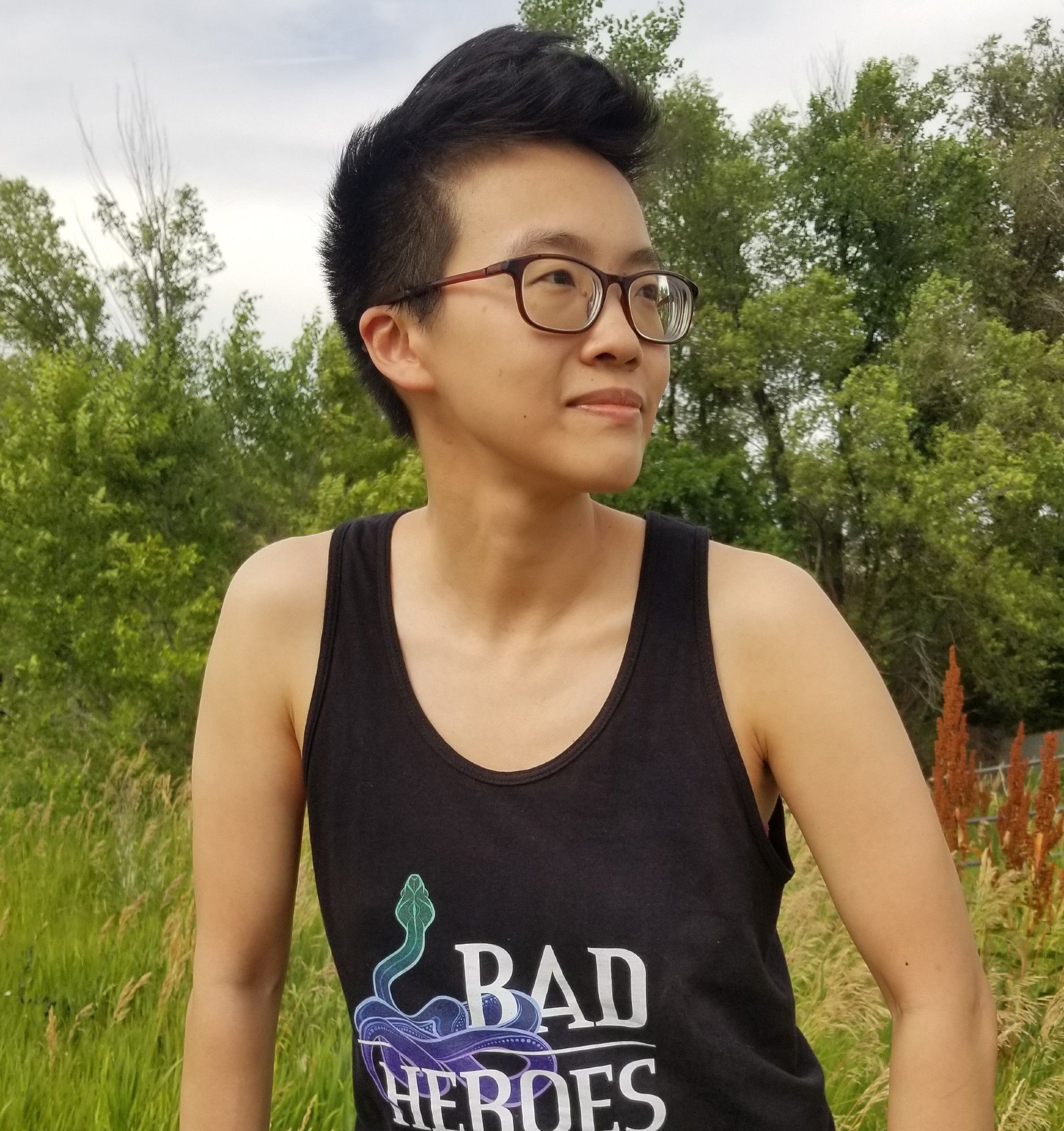 A photo of Lian looking to the right of the camera, with trees and tall grasses behind her. She has short black hair and is wearing glasses and a black tank top with the Bad Heroes logo on the front.