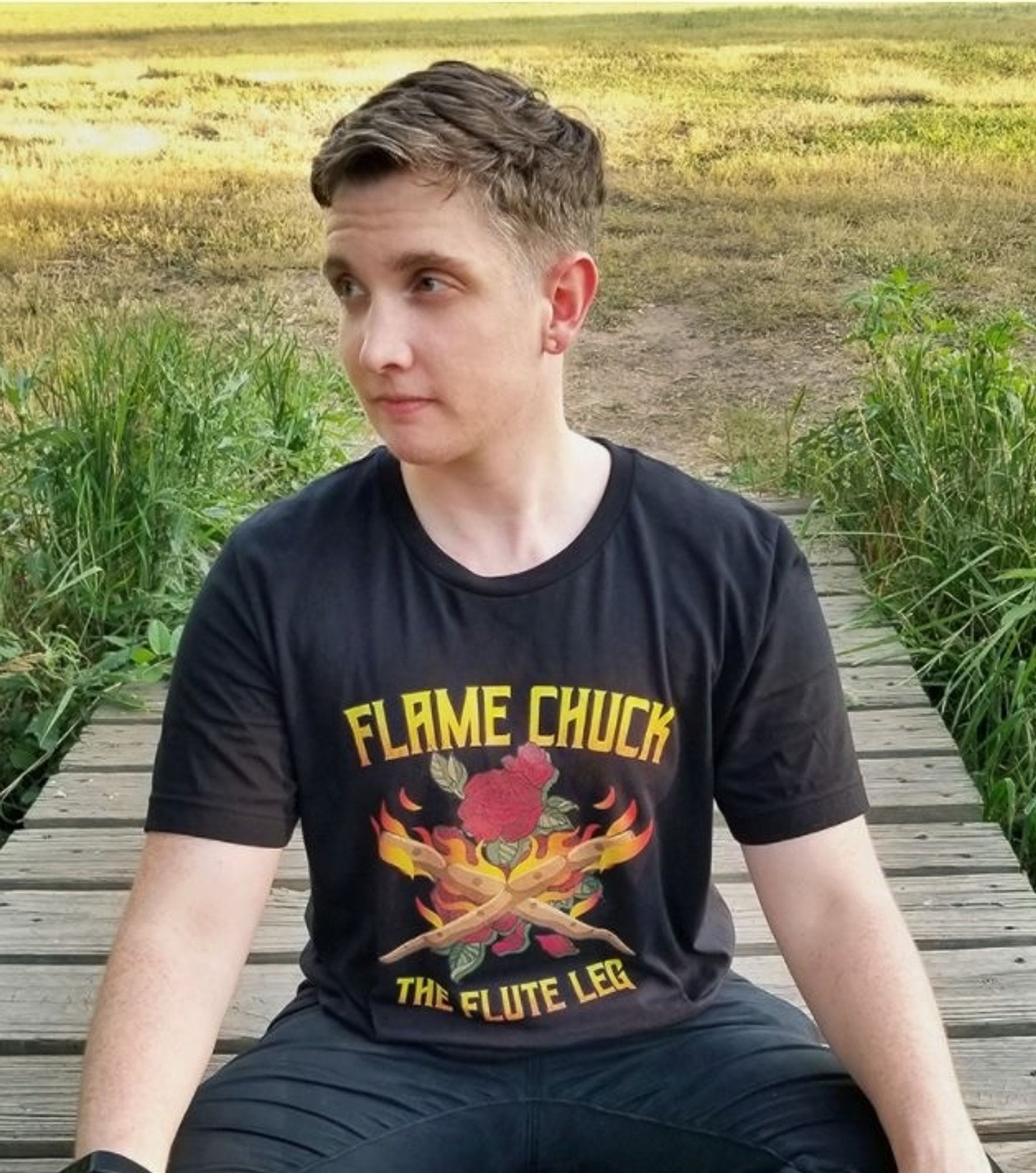 A photo of Rev sitting crosslegged and looking thoughtfully to the camera's left. He has short brown hair and is wearing dark jeans and a black t-shirt that says 'Flame Chuck the Flute Leg', a quote from the podcast.