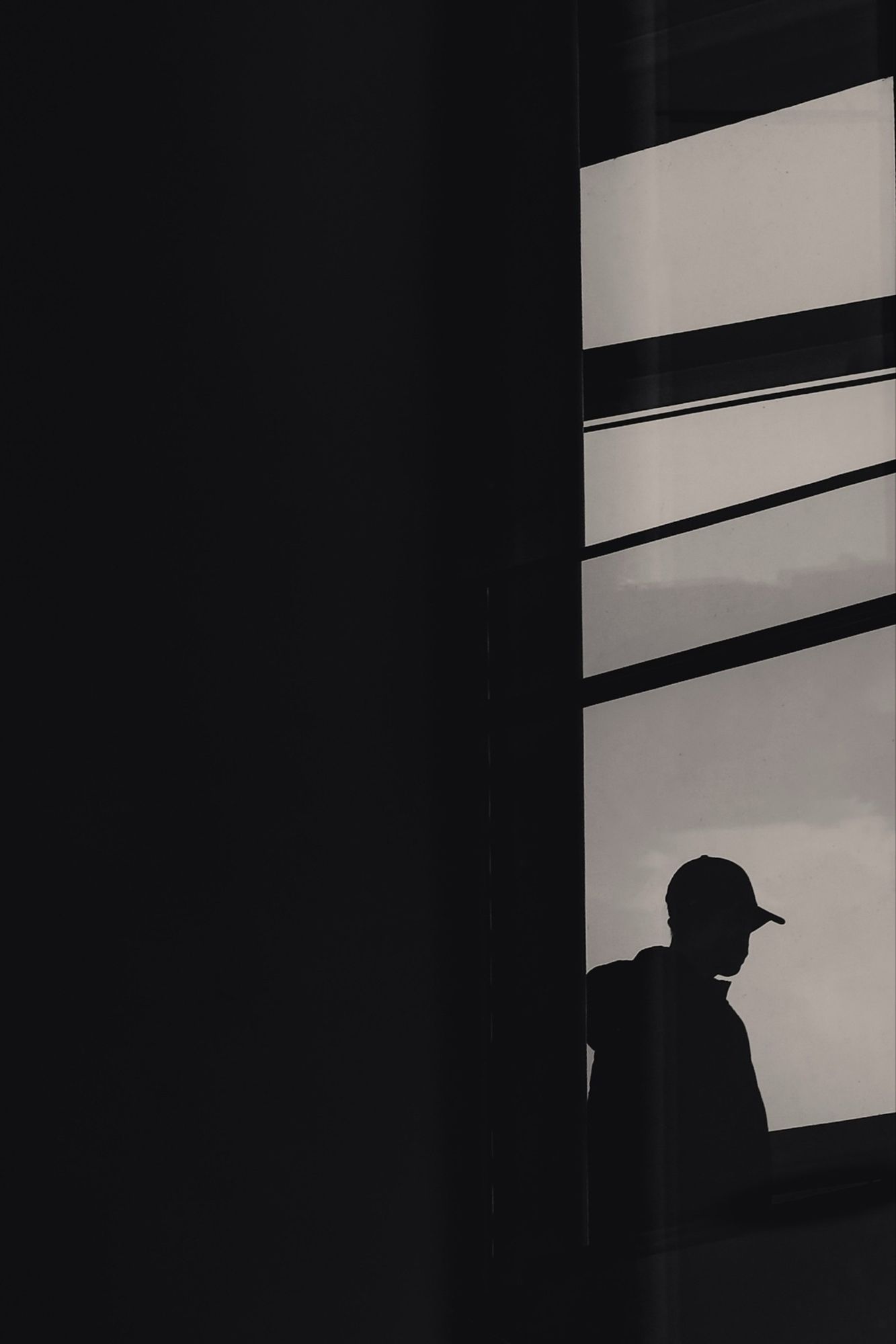 Silhouette of a person wearing a cap, standing by a window. The scene is in black and white, with strong contrasts between the dark figure and the light background.