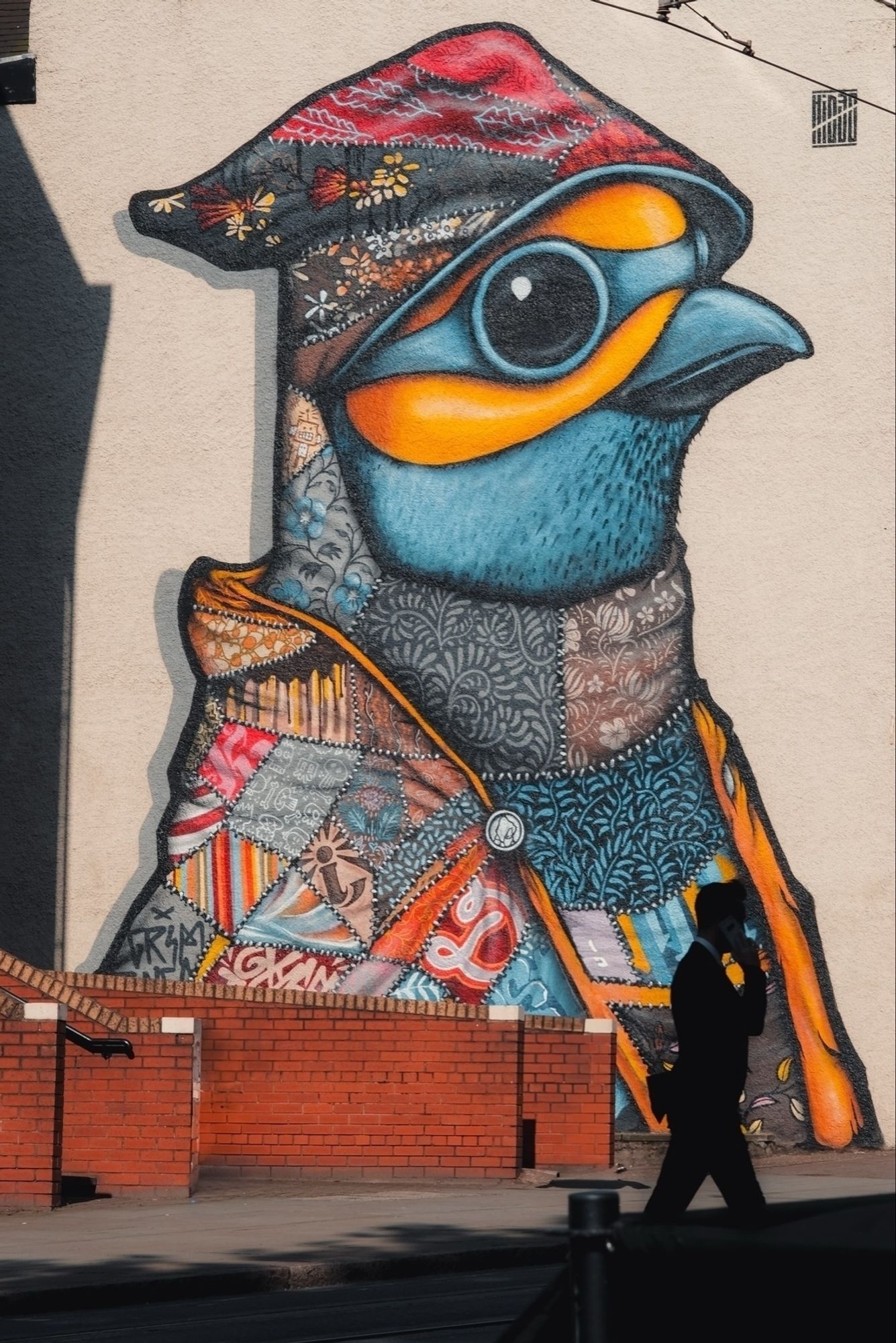 A vibrant mural of a stylized bird in patchwork clothing is painted on a wall. A silhouette of a person walking and talking on a phone is visible in the foreground. The bird features elaborate patterns and bright colors.