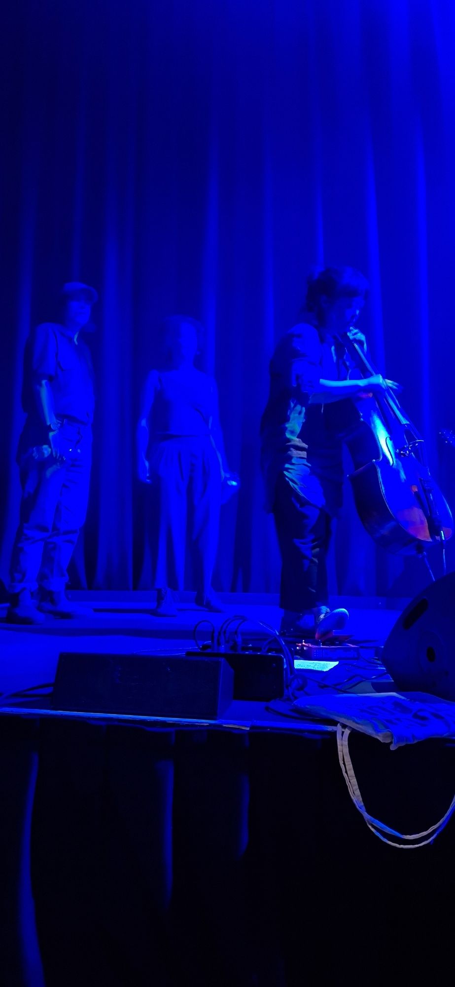 Photo of musicians. Set in blue, a cello player is tuning her cello watched by two other people.