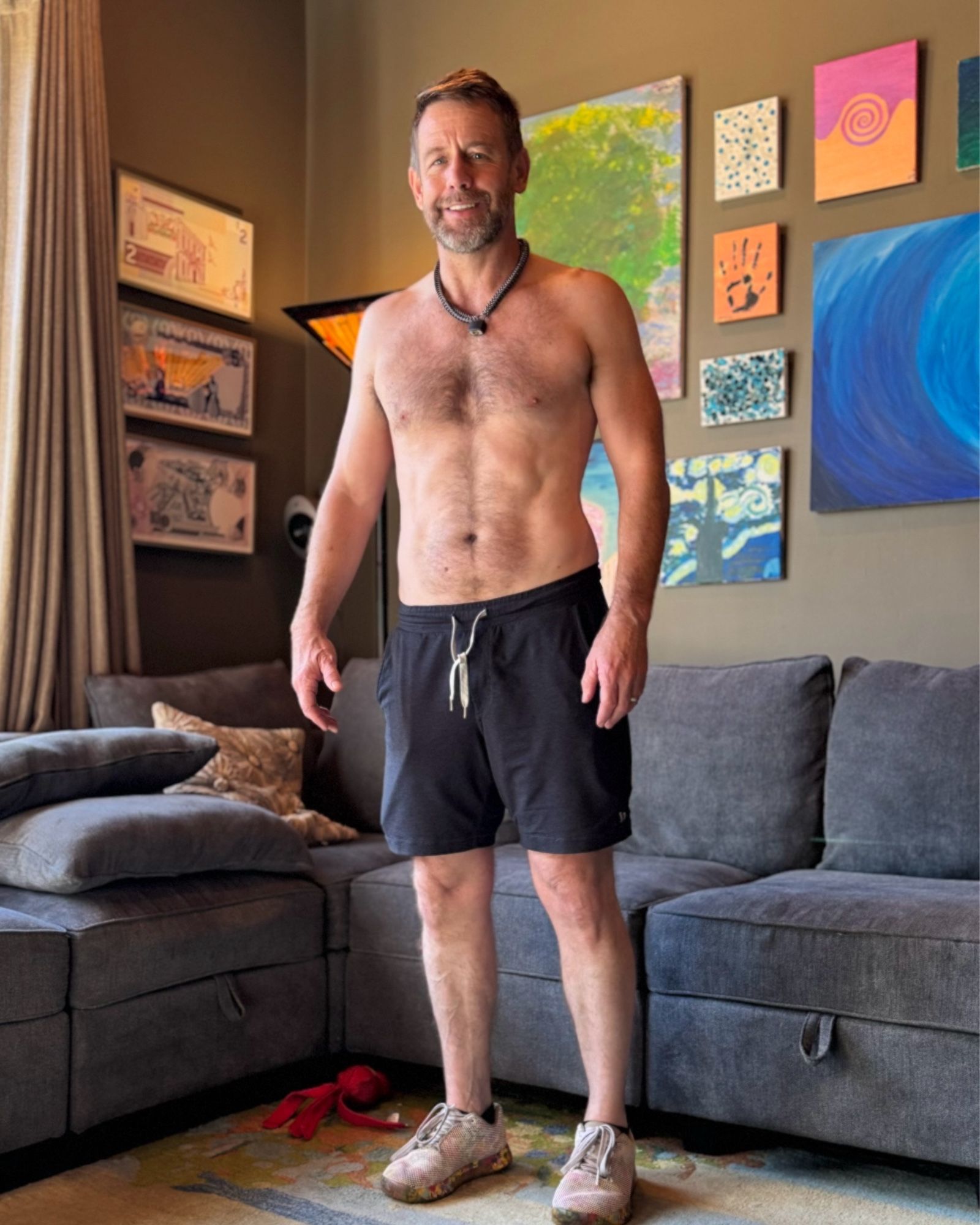 Me in untied shorts and no shirt standing in front of a blue sectional couch that has not been molwsted by JD Vance. The walls are a lovely olive shade and covered with lovely artwork.