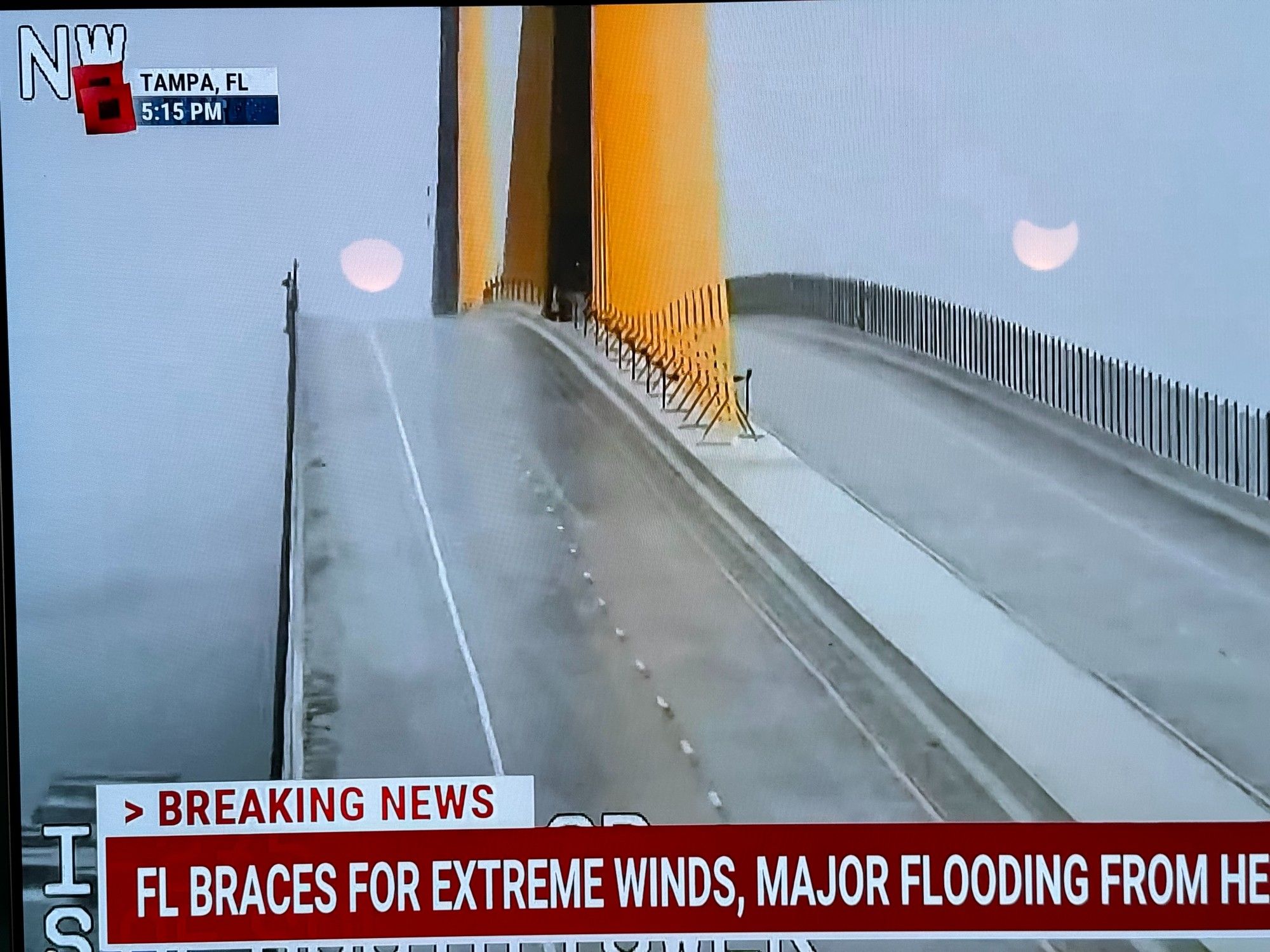 Tampa FL bridge empty of traffic and being pounded by heavy waves and surf in gray skies with TV chron overlaid talking of major FL winds and flooding from Hurricane Helene. The bridge has two lanes in each direction in gray concrete with bright yellow suspension spans in the middle.