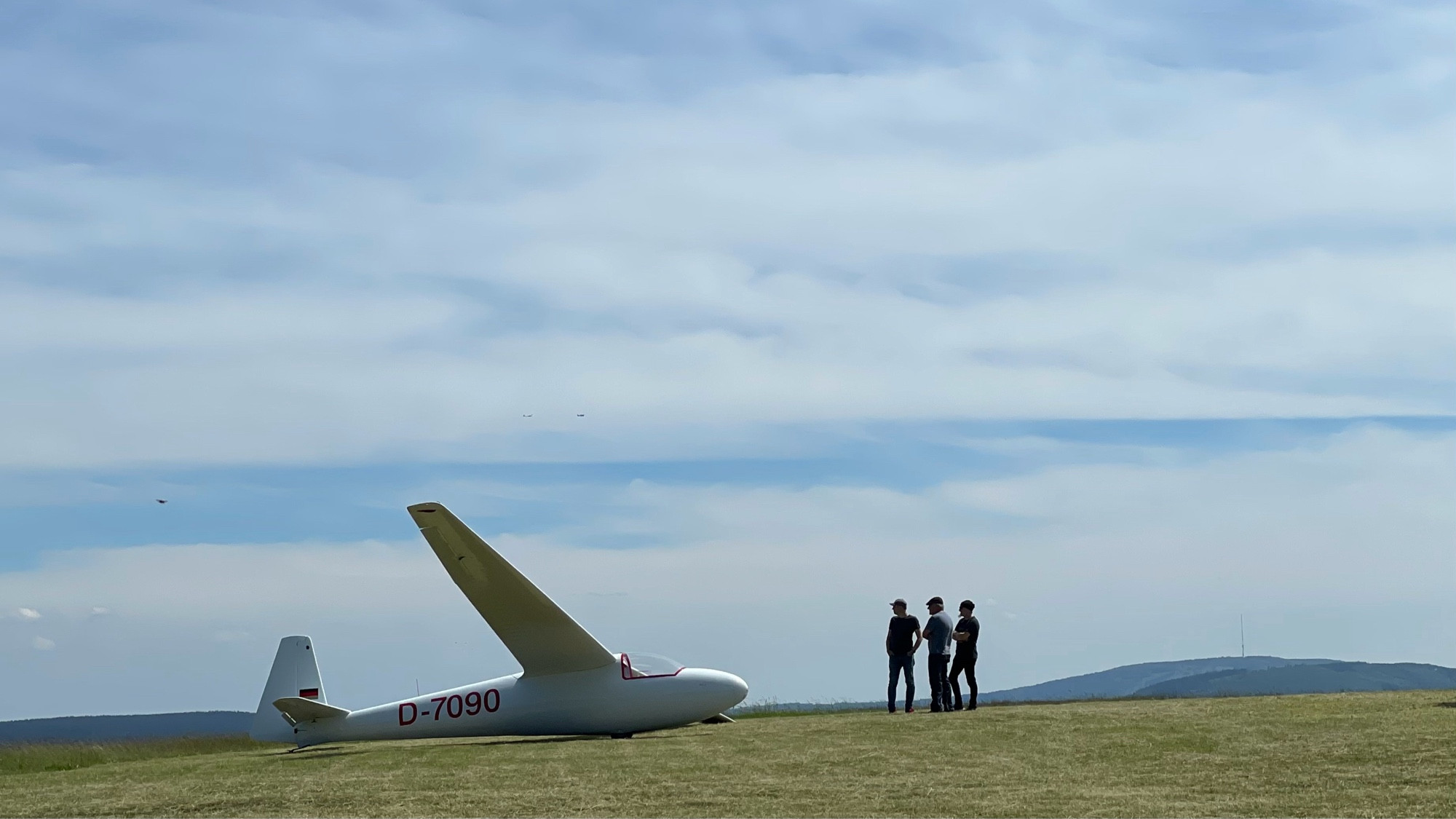 Ein Segelflugzeug am Boden. Rechts daneben drei Männer, die es betrachten. Es ist ein sonniger Tag.